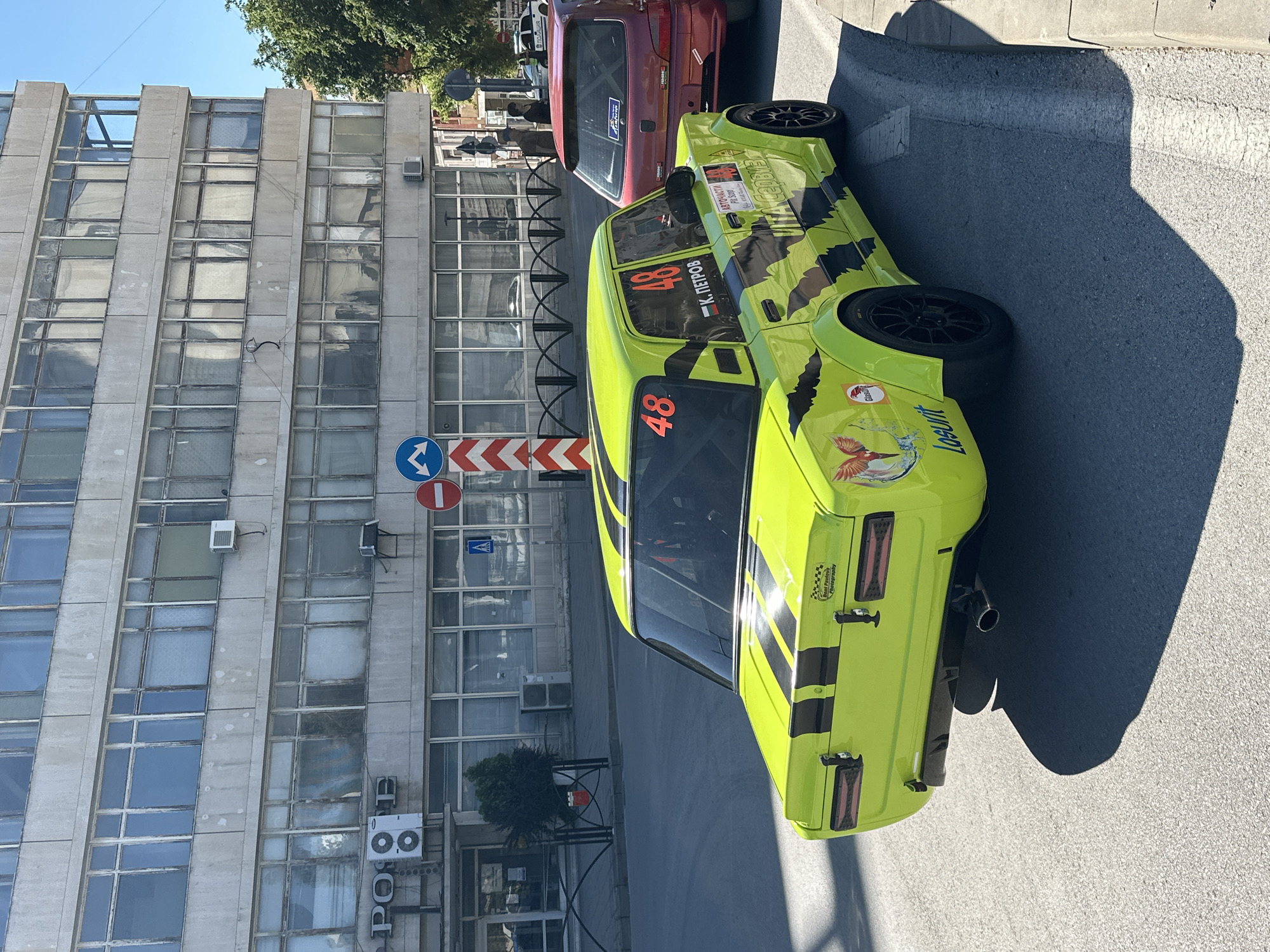 A bright yellow, modified sedan with black racing stripes and camouflage-style decals is parked on an asphalt road in the city of Gorna Oryahovitsa, Bulgaria. The car, identified by the number "48" and the name "К. ПЕТРОВ" on its side, has a racing roll cage visible through the windows. Next to it, a red hatchback is also parked. In the background, a multi-story building with a facade of concrete panels and numerous windows stands tall. A traffic sign with a blue arrow and a red circle indicating a prohibition is visible on a pole. The scene is illuminated by bright sunlight, casting distinct shadows on the road and sidewalk. The environment suggests an urban setting, likely during daytime.
