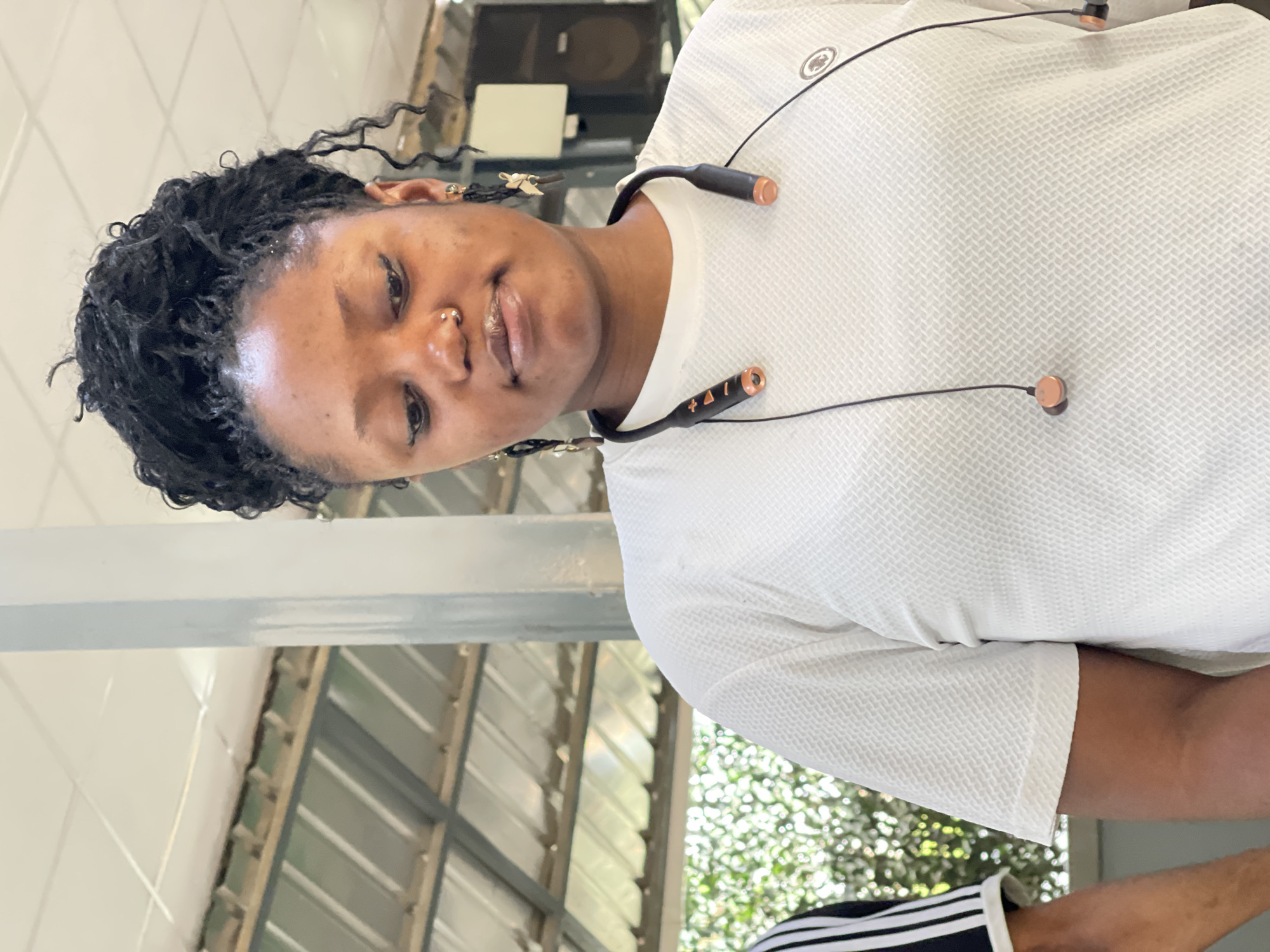 A young woman with dark skin and dark, micro-locs or braided hair is captured in a portrait-style image, rotated 90 degrees clockwise. She presents a gentle, slight smile, and has a small silver hoop nose ring in her left nostril. She wears a white t-shirt with a textured, possibly waffle-knit, pattern. Around her neck is a pair of black wireless earphones featuring copper-colored accents on the earbuds and neckband. One side of the earphone neckband displays the symbols "+ Δ !". A small, circular emblem is visible on the upper right side of her shirt.
The background suggests a well-lit, possibly semi-outdoor or open indoor setting. There are light-colored architectural elements, including a metallic beam and what appears to be a window or series of louvers. Through these, bright green foliage and natural light are visible, indicating it is daytime. The scene conveys a relaxed and casual atmosphere, consistent with a location like Abuja, Nigeria.