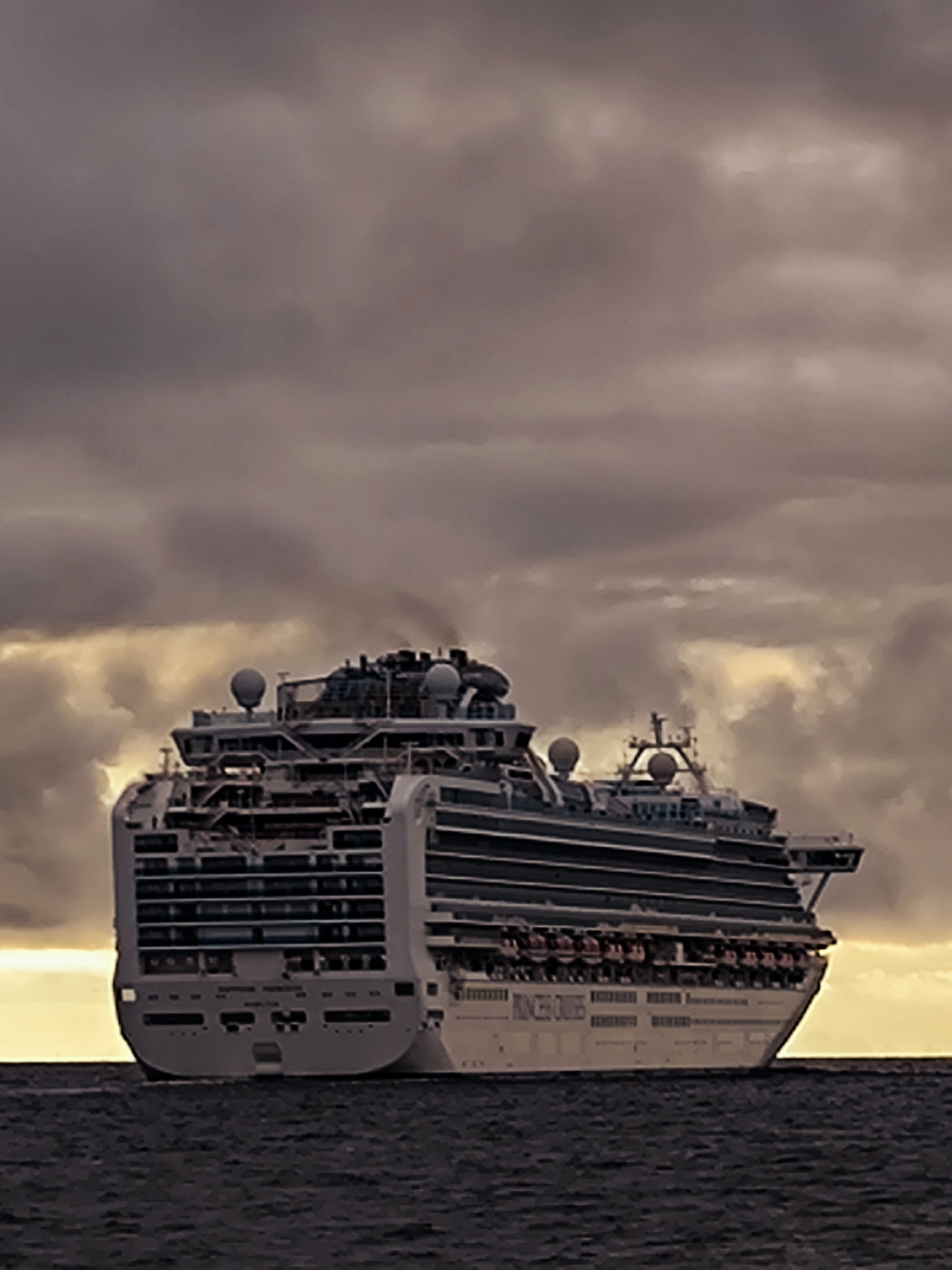 A large cruise ship, identified by the text "PRINCESS CRUISES" on its hull, is sailing on dark, choppy water under a cloudy, overcast sky. The ship is white with multiple decks, featuring numerous windows and balconies. Lifeboats are visible along the sides. The setting sun casts a warm glow through a break in the clouds towards the horizon. The scene is devoid of any people or specific landmarks that would identify it as being in Riga, Latvia.