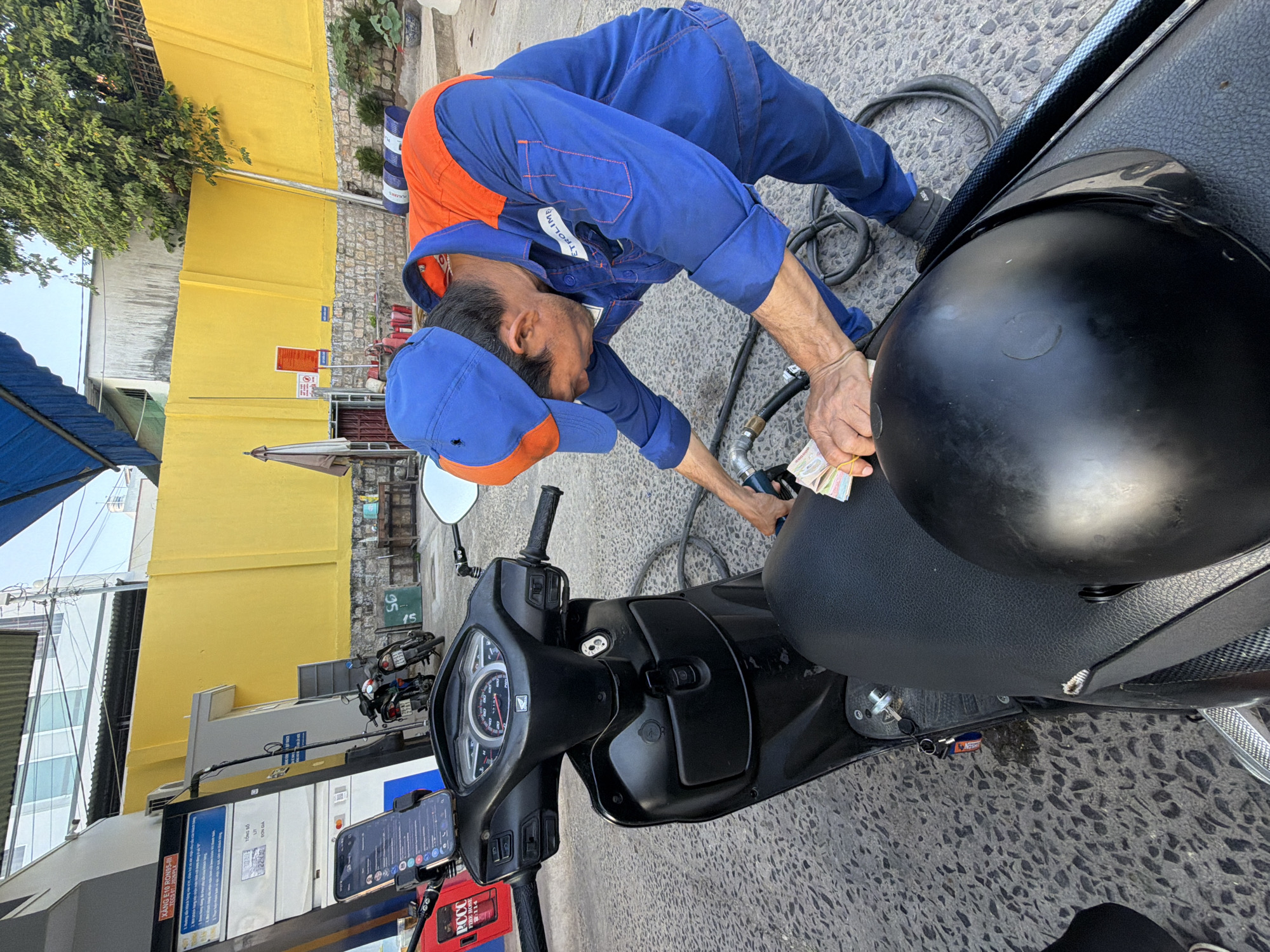 A man in a blue and orange uniform is refueling a black scooter at a gas station. He is holding a nozzle in one hand and a wad of Vietnamese currency in the other. A black helmet rests on the scooter's seat. The gas station has a yellow wall in the background. The scene appears to be outdoors, on a paved surface.