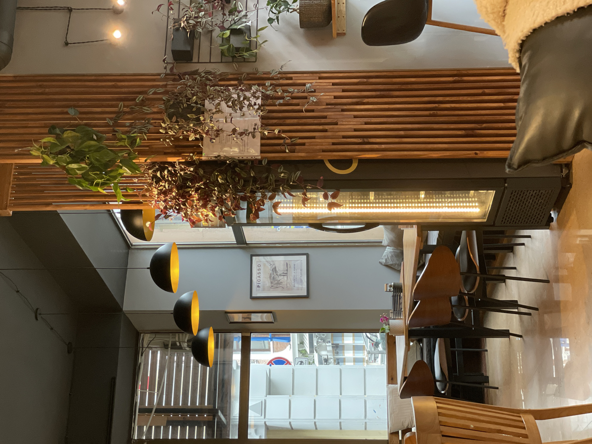 The image shows the interior of a cafe or restaurant, possibly in Batumi, Georgia, based on the "Picasso" framed print on the wall, which could indicate a connection to art or a namesake theme. The establishment appears to be located on a street with a visible view of a crosswalk, buildings, and possible street activity through the window. The scene consists of tables and chairs arranged in the dining area, with plants hanging from the ceiling and wall, creating a decorative atmosphere. The lighting consists of a row of light fixtures with black shades and a warm, yellow glow. There is also a wooden slat wall design element and a heating unit suspended above the tables. The presence of a chair and the corner of a leather object suggests that the photo was taken from a seated position. No people are visible in the frame.