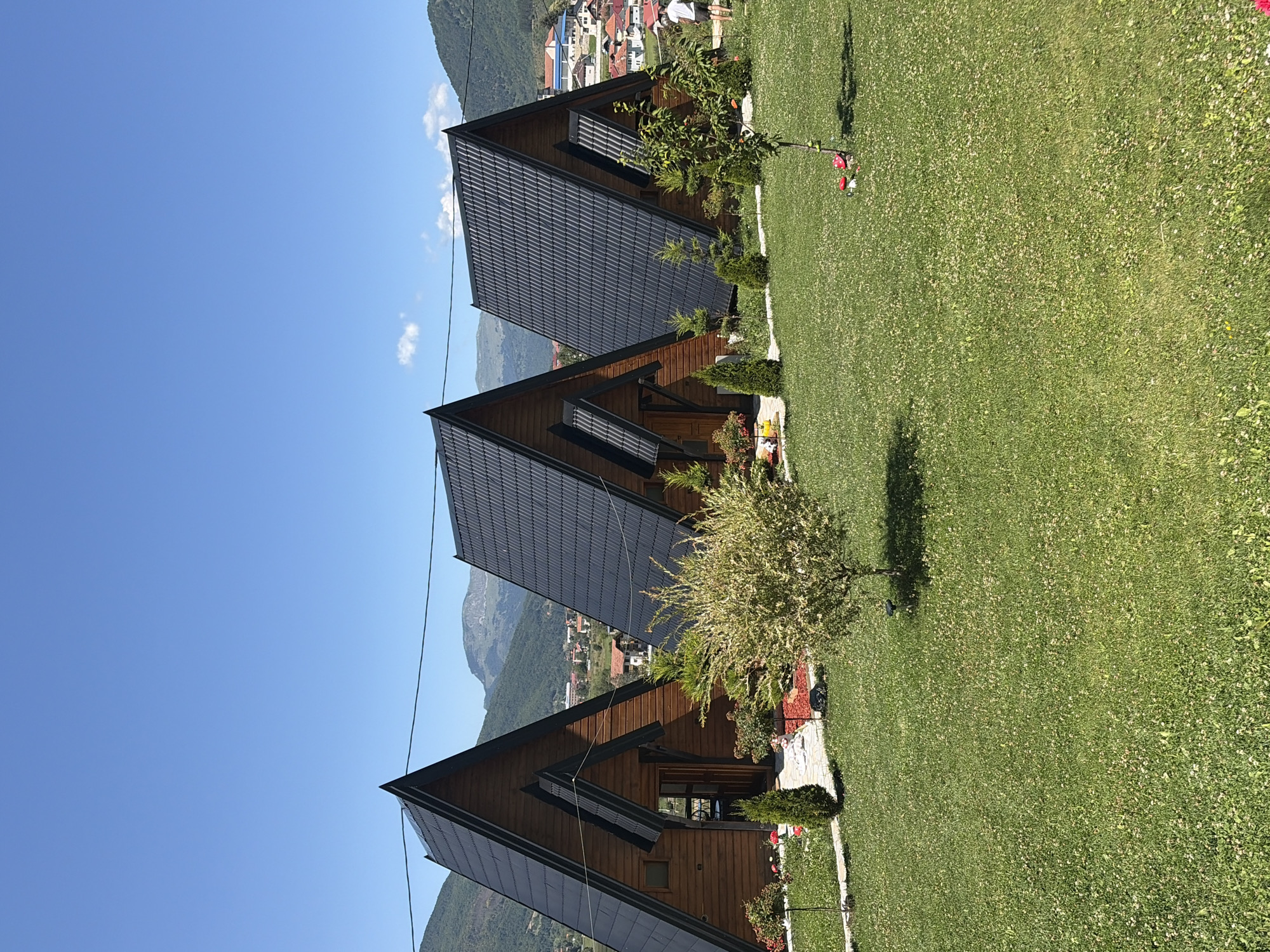 The image depicts a scenic view of three triangular wooden cabins with dark gray roofs, set against a backdrop of lush green hills and a clear blue sky. The cabins are arranged in a row, with manicured grass and small trees in the foreground. In the distance, a town with red-tiled roofs is visible nestled among the hills, suggesting the location is Plav, Montenegro. The scene is peaceful and idyllic, likely a resort or tourist accommodation. A person is visible in the background, near the third cabin, walking away from the camera. The overall impression is one of a tranquil, natural setting with modern amenities.