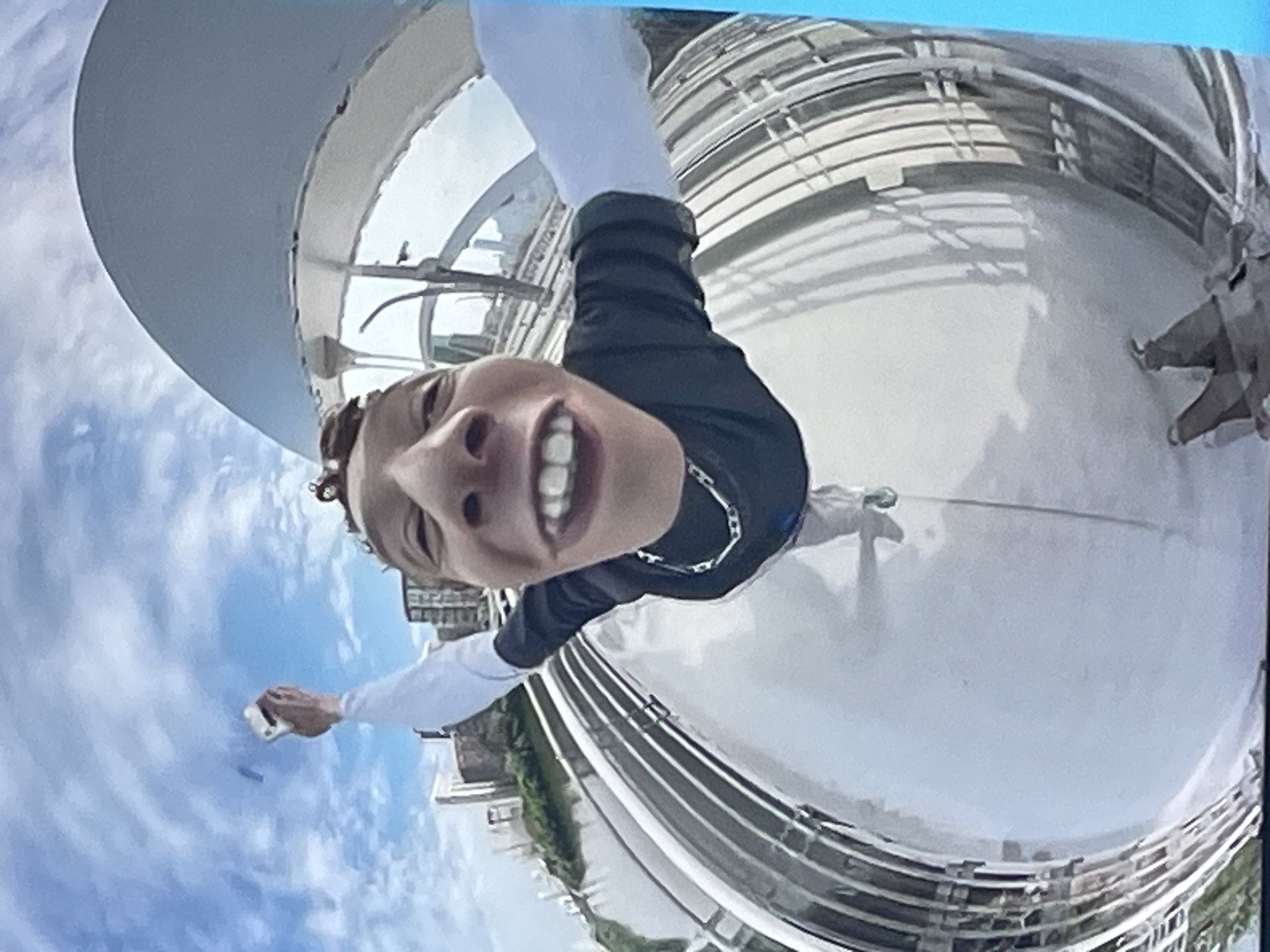 The image is a distorted, fisheye lens photograph of a person holding up a white object, likely a phone, to take a selfie. The person is wearing a black t-shirt with a white long-sleeved shirt underneath, and a silver chain necklace. They are smiling widely, with their face taking up a large portion of the frame due to the wide-angle lens. The background appears to be an outdoor urban setting with a river or body of water to the left, and buildings in the distance. A large, curved, white structure dominates the upper right portion of the image. The perspective is from a low angle, looking up at the person. The image does not depict the city of Zhukovo, Russia.