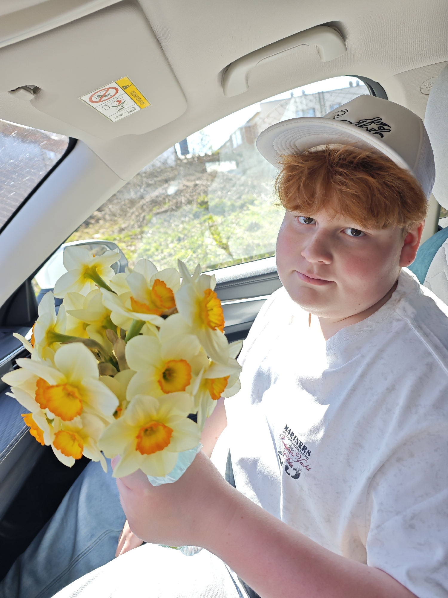 A person with red hair and a white baseball cap is sitting in a car, holding a bouquet of yellow and white daffodils. The person is wearing a white t-shirt with a nautical-themed graphic. The car's interior is light gray, and a window shows a blurry outdoor scene. The location appears to be inside a car, and no specific indicators suggest the city of Фоки, Russia.