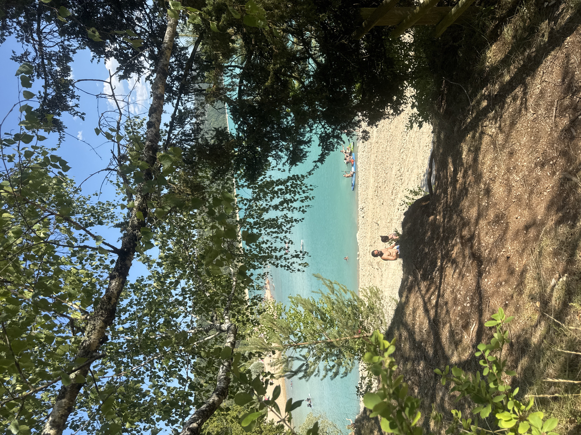 The image captures a sunlit scene at the Lac de Vouglans in Maisod, France. In the foreground, a sandy slope is dotted with scattered pebbles and dried grass, with shadows cast by overhanging trees. On the slope, two individuals, one a shirtless male and the other a person wearing a dark top, are seated.
Beyond the slope, the turquoise waters of the lake stretch out, dotted with several people. Some are swimming, while others are engaged in water sports, including paddleboarding. A group of individuals is visible on paddleboards near the shore. The far bank of the lake is lined with trees and hills. The sky above is a clear blue with a few wispy clouds. The overall impression is one of a leisurely summer day at a popular lakeside recreational area.