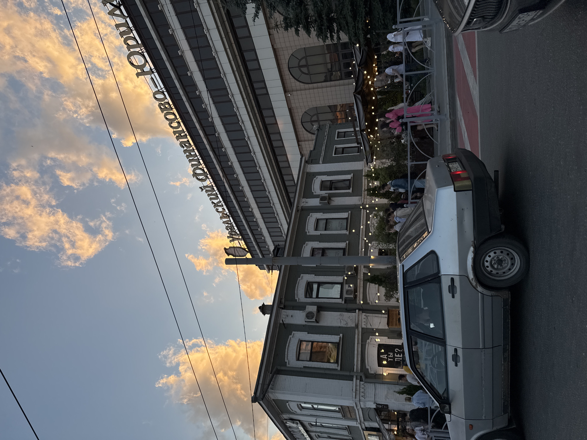 The image shows a street scene in Yekaterinburg, Russia, likely in the evening, with dramatic clouds in the sky. In the foreground, a silver car is parked on the right side of the road. Further back, a two-story building with a green facade and white trim stands to the left, featuring multiple windows and an air conditioning unit. A sign above one of the entrances reads "А ты где?" (Where are you?).
Behind the green building, a larger, modern building with a facade of glass and beige stone dominates the background. Large, arched windows are visible on the lower floors of this building. The text "УРАЛЬСКИЙ ФИНАНСОВО-ЮРИД" is prominently displayed in large, white lettering across the top of this modern structure.
Several people are visible in the scene, some standing near the green building, others walking along a sidewalk or pathway separated by a metal railing. String lights are strung along the front of the green building and under awnings, suggesting an outdoor dining or seating area. The sky is filled with large, fluffy clouds illuminated by warm, orange-yellow light, indicating either sunrise or sunset. Electrical wires are strung across the upper portion of the frame.