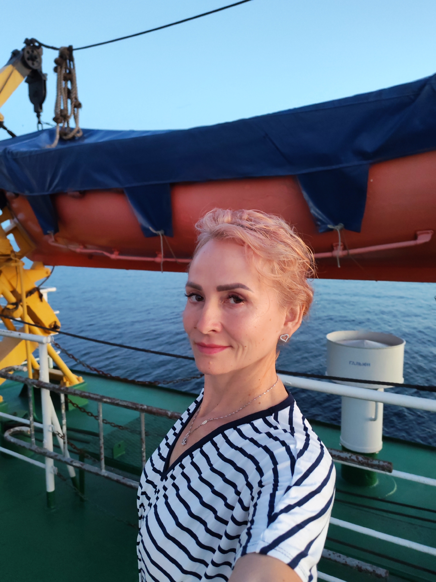 An exterior, waist-up, eye-level medium shot captures a woman with short, pink-tinted hair and a light-hearted expression posing for a selfie on the deck of a ship. She is wearing a black and white striped v-neck top and a silver heart-shaped earring. Behind her, a bright orange life raft is secured to the side of the ship, with a dark blue cover partially obscuring it. The vast, calm blue sea stretches out behind the ship. A yellow crane arm extends from the left side of the frame, and a white vent with Russian Cyrillic text reading "ГАЛЬЮН" (likely "head," referring to a toilet vent) is visible on the right. The scene appears to be set in the city of Shal'skii, Russia, based on the provided context.