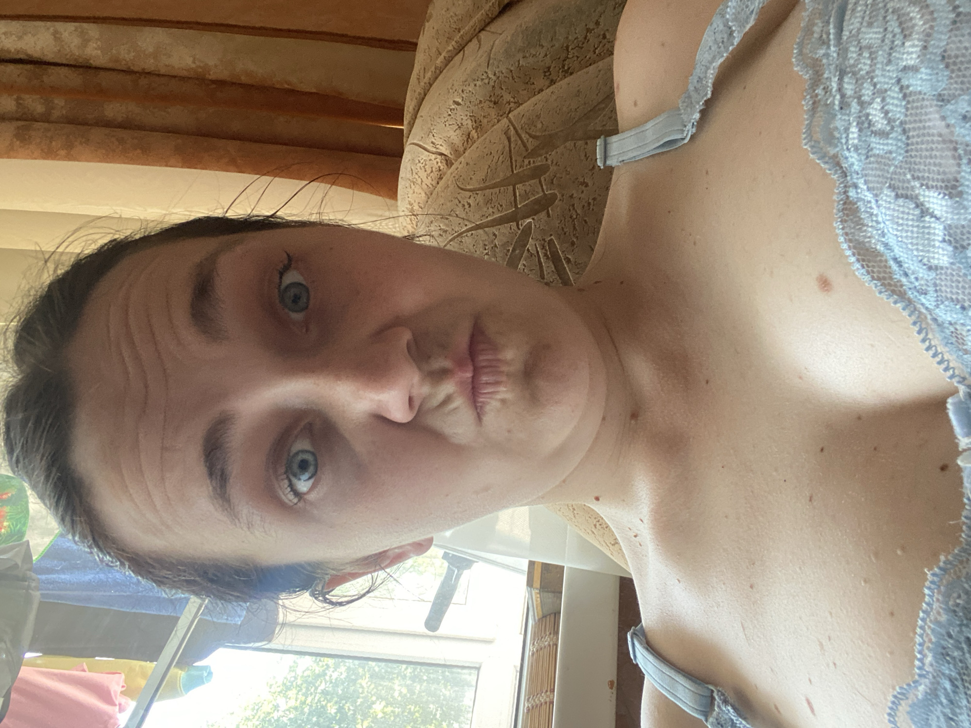 A woman is captured in a close-up selfie. She has her hair pulled back, revealing her forehead with visible wrinkles. Her eyes are wide open and blue, with her eyebrows slightly raised. Her mouth is pursed, with lips pushed forward in a pouty expression. She is wearing a light blue lace bra. The background includes a window with blinds and curtains, as well as a patterned armchair. The lighting suggests it is daytime.