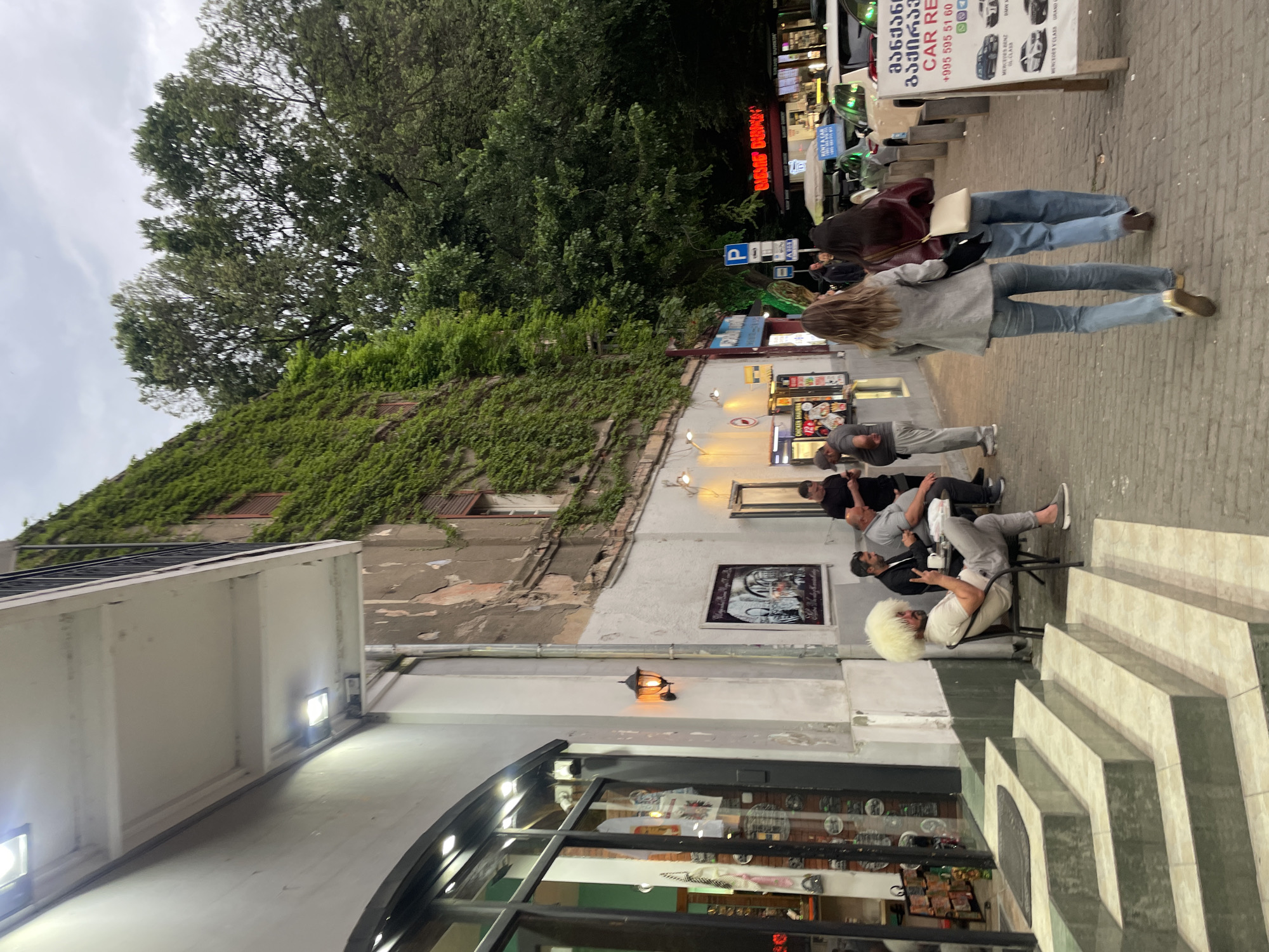 The image depicts a street scene in Tbilisi, Georgia. People are present, some walking along the sidewalk, while others are seated on chairs outside a building. A man in a white wool hat is seated. The building is partially covered in green ivy. Various signs are visible, including advertisements for car rentals. Street lamps provide illumination. A shop with a curved window display is at the bottom of the frame. The environment appears to be a mix of commercial and residential spaces.