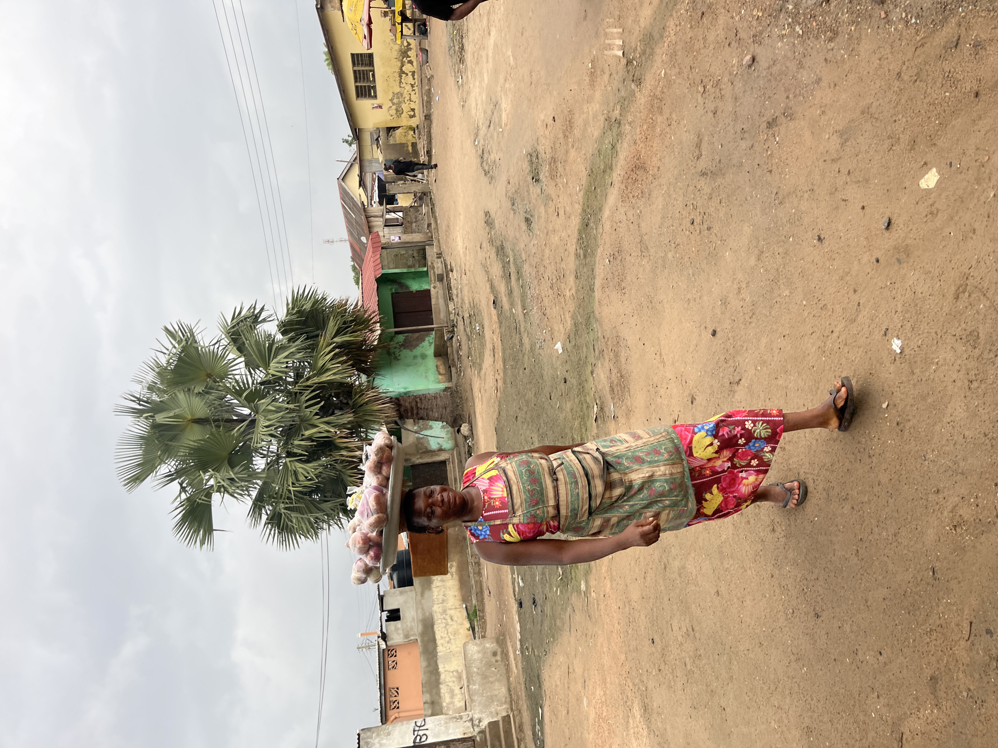 A woman walks from left to right in a dusty, unpaved street in a neighborhood in Accra, Ghana. She is carrying a large round tray of goods, likely produce, balanced on her head. The tray appears to contain an assortment of red and white items, some of which are wrapped in plastic. She wears a brightly colored floral print dress and an apron. Her feet are shod in dark flip-flops. Behind her, to the left, is a low, cream-colored building with some graffiti. To her right, a large palm tree is visible. Further in the background are various low-rise buildings with corrugated metal roofs and some yellow structures. The sky is overcast. The scene depicts a common mode of commerce and transportation in a Ghanaian urban environment.
