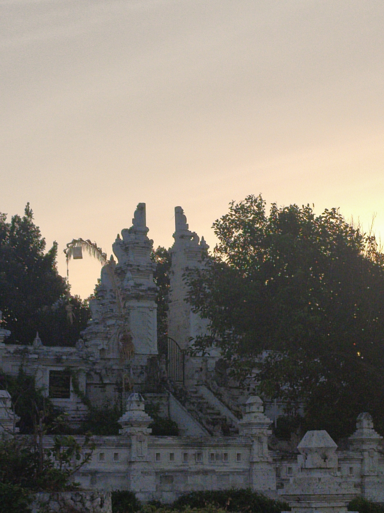 A detailed, forensic, matter-of-fact description of the scene, including essential details about people, subjects, objects, actions, interactions, and the environment, is provided below.
The image depicts an exterior view of what appears to be a temple or a cultural site, likely in Kuta Selatan, Indonesia, given the architectural style. The sky is a pale, hazy orange, indicative of either sunrise or sunset. In the foreground, a complex of white, ornately carved stone structures dominates the scene. These structures feature multiple tiers, pilasters, and decorative finials. A prominent element on the left is a tall, multi-layered gateway with intricate carvings. To its right, a similar, slightly smaller gateway stands. Between these structures, a dense, dark green tree is visible, its foliage obscuring some of the background. Further to the right, another large tree with dark green leaves is present, partially silhouetted against the sky.
Hanging from a structure in the mid-ground, between the two main gateway structures, is a traditional Balinese Penjor, a decorative bamboo pole adorned with leaves and a small white cloth, which is often seen during religious ceremonies. A metal grate, likely a gate, is visible behind the structures on the right, leading up a set of stairs. The overall condition of the stone structures appears weathered, with some areas showing signs of age and possibly neglect, overgrown with some vegetation. The lighting is soft and diffused, casting gentle shadows. No people are visible in the frame. The environment appears to be a tropical or subtropical setting, suggested by the lush foliage.