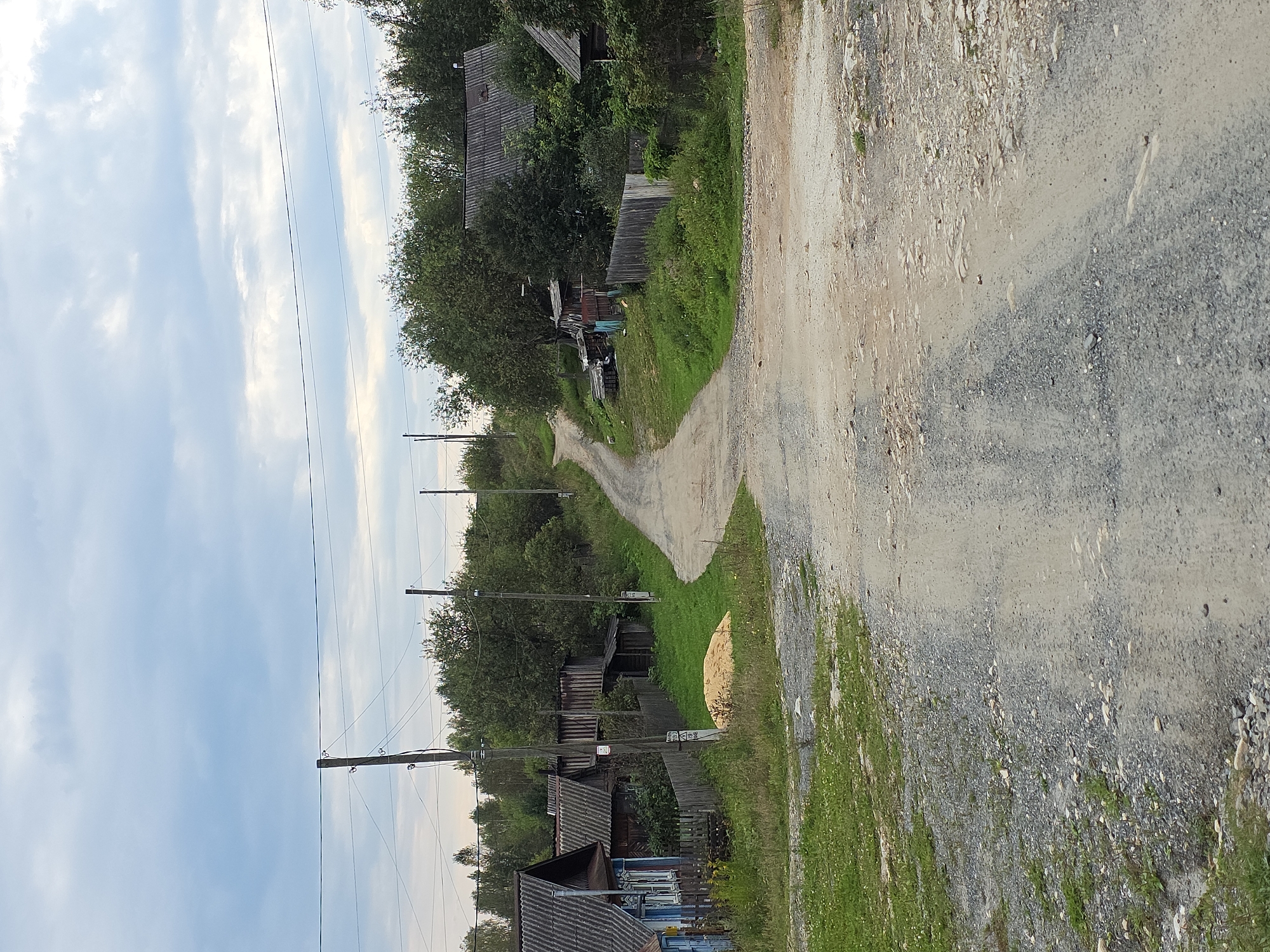 The image depicts a rural dirt road winding through a village under a cloudy sky. The road is unpaved, with a mix of gravel and dirt, and appears to be somewhat uneven. On either side of the road are wooden houses with corrugated metal roofs, typical of rural Russian architecture. Some houses have fences and gardens. Trees and green vegetation surround the houses and line the road. Several utility poles with wires are visible along the road. There is a pile of what appears to be sawdust or sand on the side of the road near one of the houses. The overall impression is of a quiet, somewhat rustic village setting. No people are visible in the image. The scene is consistent with a location in a rural area such as Magnitka, Russia, characterized by its simple infrastructure and traditional housing.