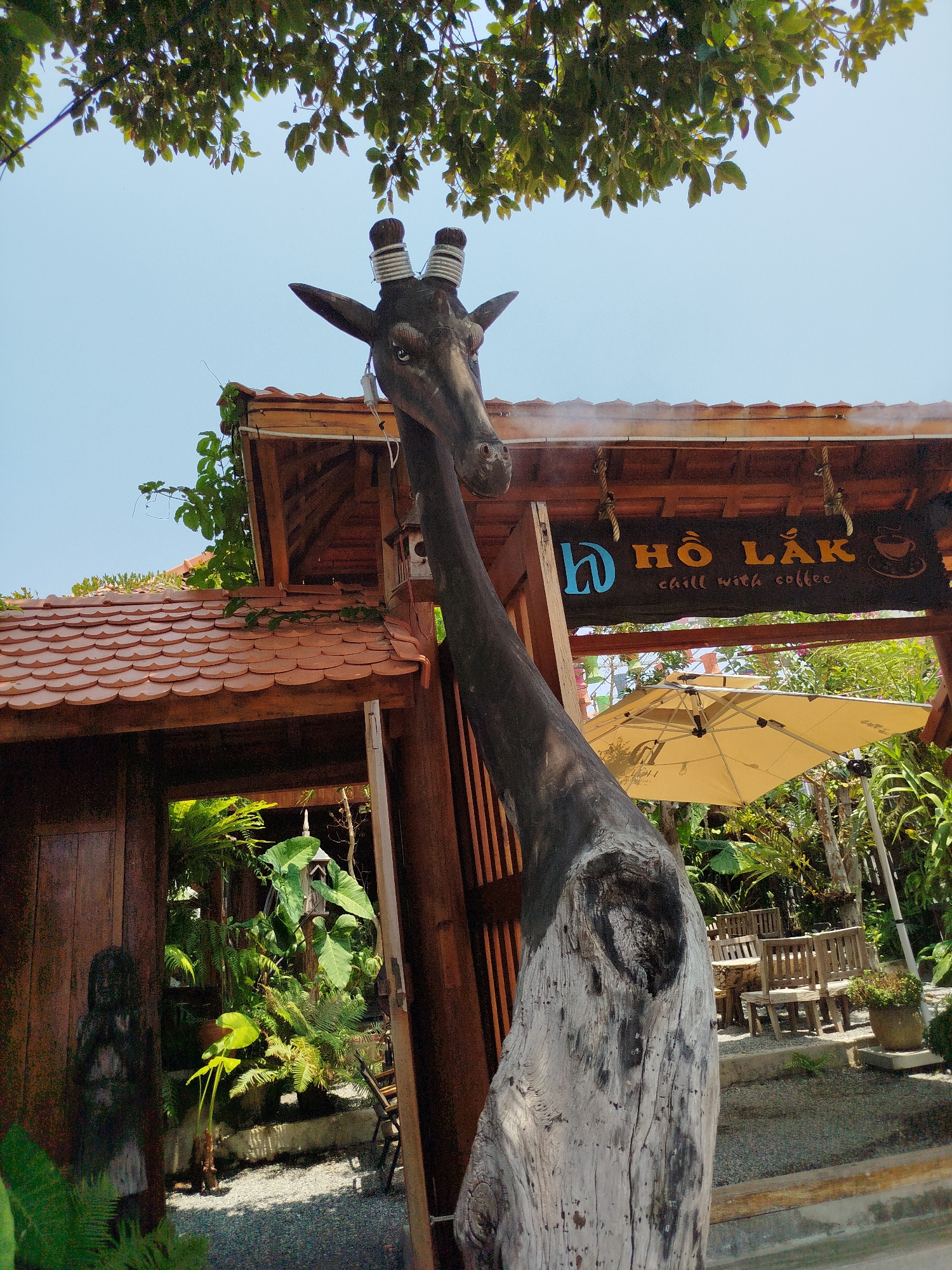 The image shows a wooden giraffe sculpture in front of a cafe named "Hồ Lắc chill with coffee". The cafe is located in Ho Chi Minh City, Vietnam. The sculpture is made of dark wood for the head and neck, while the body is made of light wood. The cafe has a sign with its name on it, as well as tables and chairs for customers. There is an umbrella over the tables. The environment appears tropical, with various plants and greenery. A small wooden structure is visible on the left side of the image.