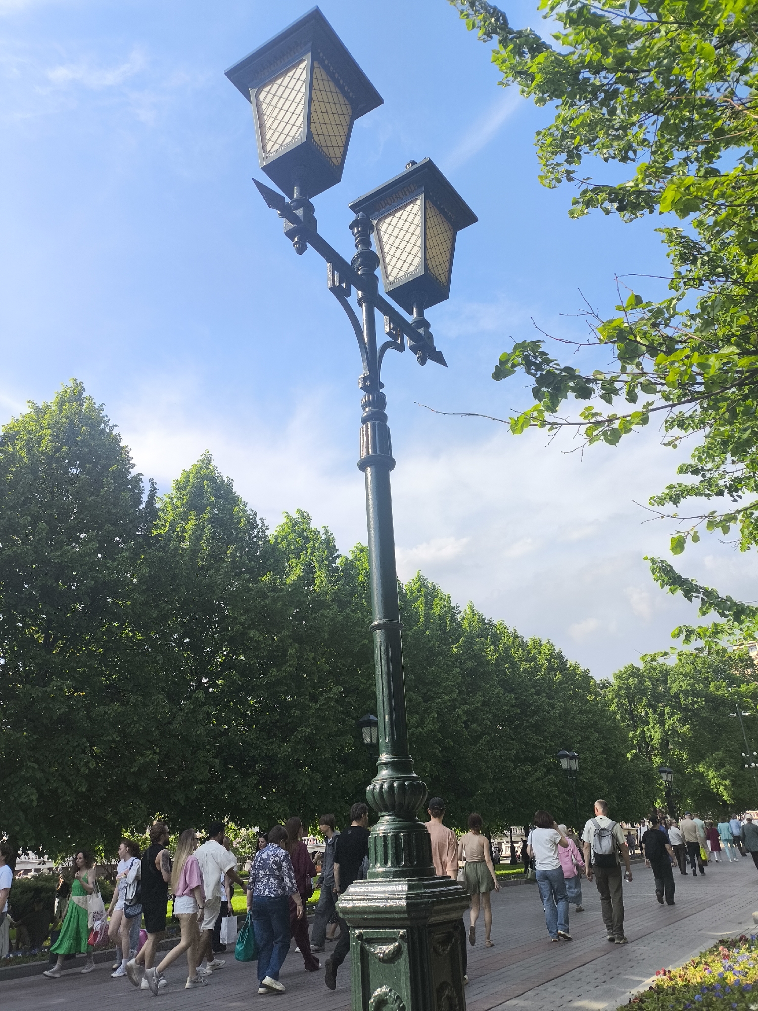 The image shows a street scene in Moscow, Russia. The foreground features a tall, ornate street lamp with two lanterns on top. A group of people is walking along a paved path in the background, lined with trees. The sky is blue with some clouds.