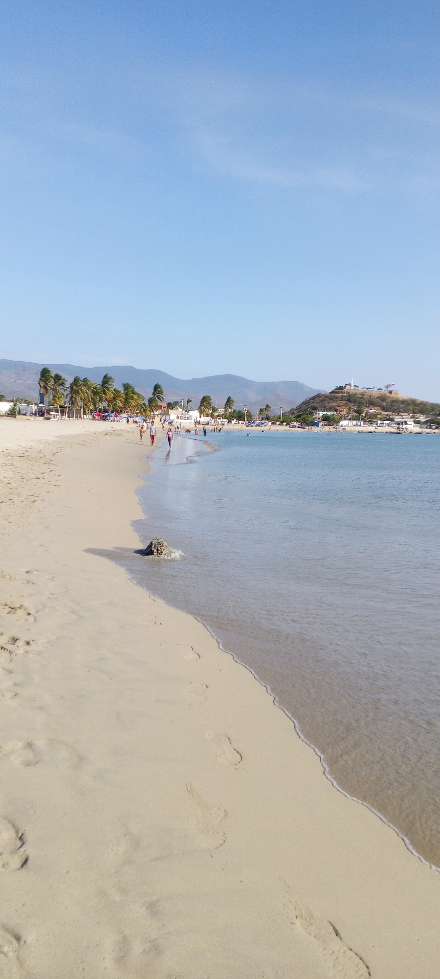 The photograph captures a coastal scene. A sandy beach stretches into a calm body of water, which merges with a clear, blue sky. In the distance, low mountains and a small, settled hilltop are visible.
Along the shoreline, several individuals can be observed walking and interacting with the water. Palm trees line the edge of the beach, suggesting a tropical or subtropical climate.
In the foreground, footprints are visible in the sand. A small, dark rock sits at the water's edge, partially submerged by the gentle waves.