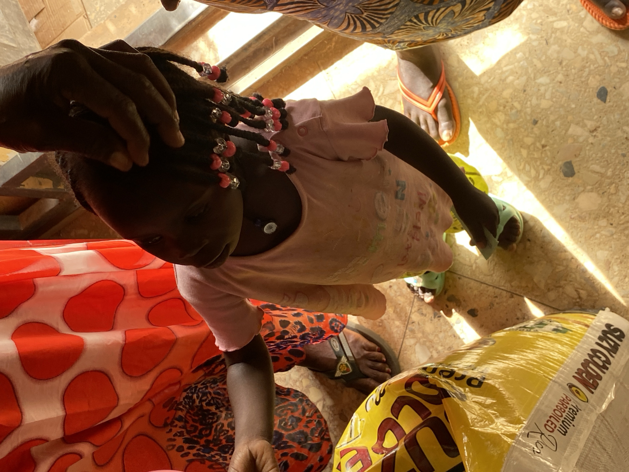 A child with braided hair and pink beads is being touched by an adult's hand. The child is wearing a light pink shirt. The child and an adult wearing a red and white patterned garment are standing near a large bag of "Suzy Golden" premium parboiled rice. Another adult in a patterned garment is standing nearby, wearing orange flip-flops. The setting appears to be indoors on a tiled floor. The location is in the city of Bukuru, Nigeria.