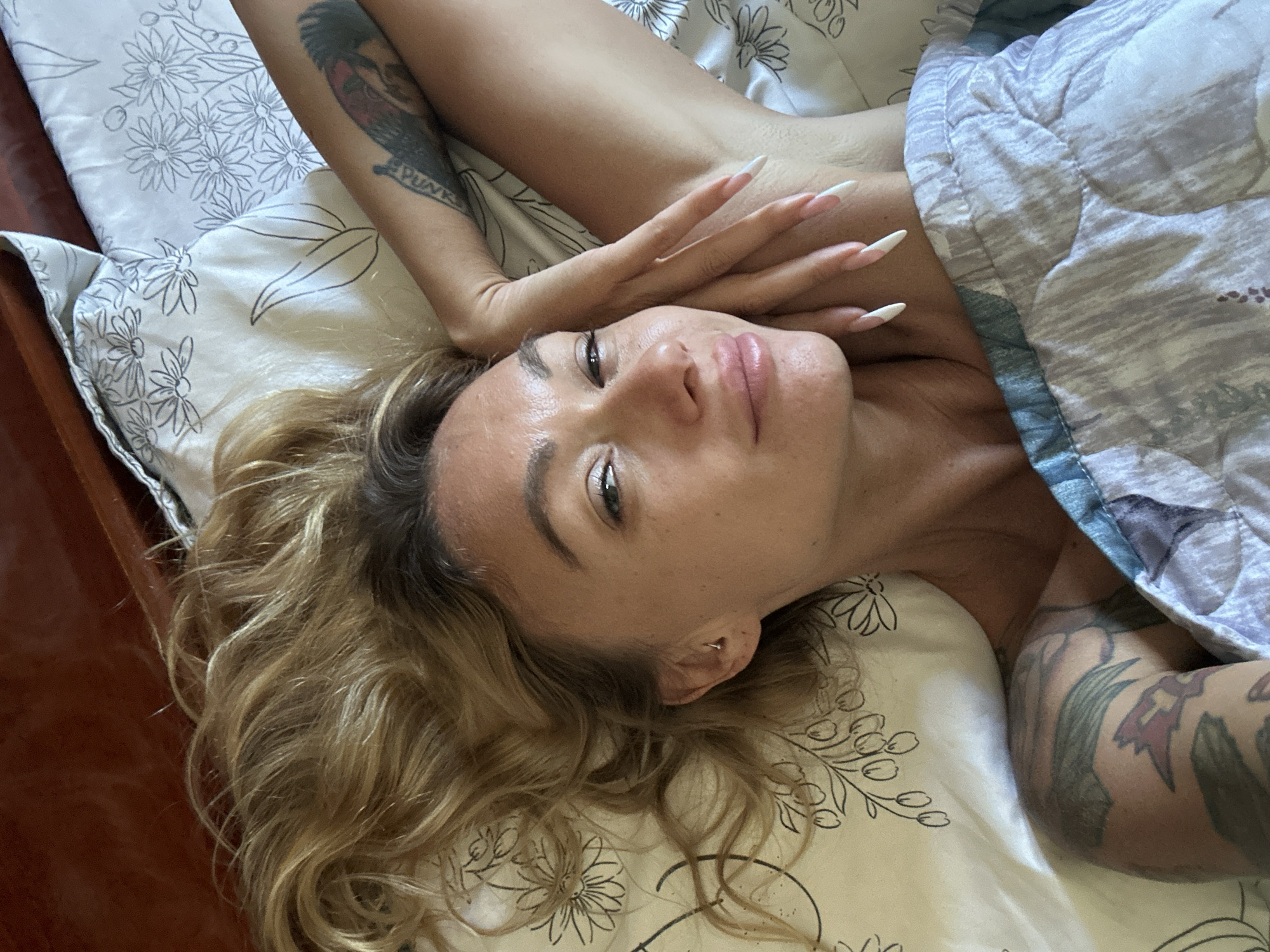 The image shows a woman lying on a bed with blonde hair, fair skin, and a slight smile. She is wearing makeup and has long, manicured nails. Tattoos are visible on her arm and shoulder. The bedding has a floral pattern. The overall scene suggests a relaxed, indoor setting, possibly a bedroom. The woman is positioned centrally in the frame, looking towards the camera.