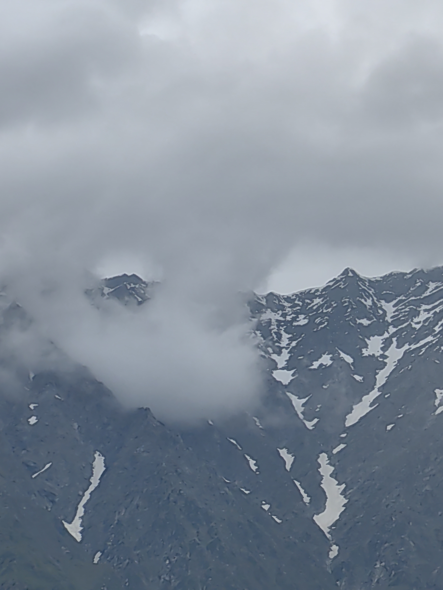 The image displays a mountainous landscape under a cloudy sky. The mountains are rugged and partially covered in snow, with patches of green vegetation visible at the lower elevations. Clouds obscure the peaks in some areas, creating a misty and ethereal atmosphere. The scene is devoid of people, man-made structures, or any discernible activity, suggesting a remote and natural environment. The overall impression is one of raw, untamed natural beauty characteristic of the Himalayan region.