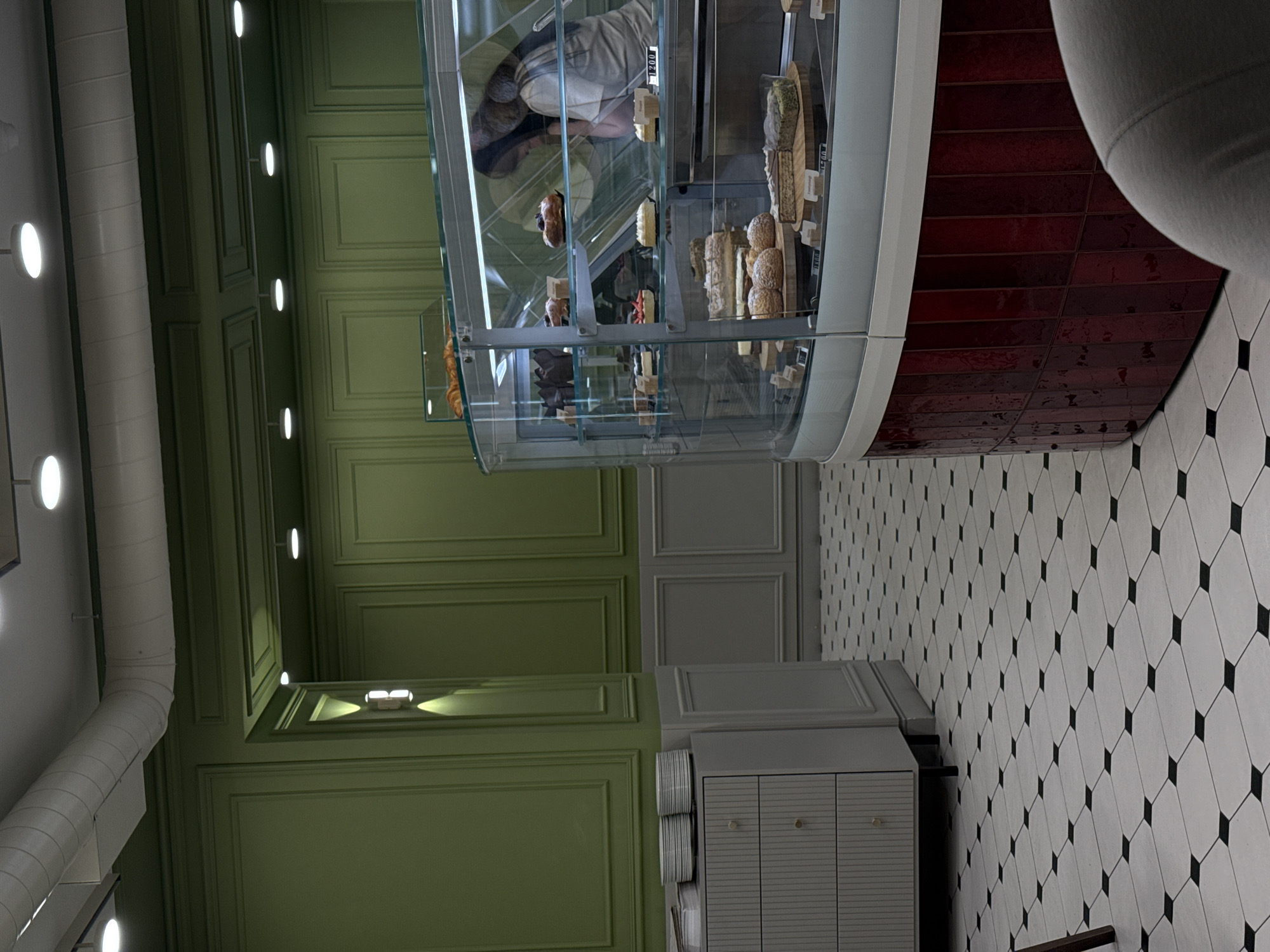 The image depicts the interior of a bakery or pastry shop in Yerevan, Armenia. The dominant features are a curved glass display case filled with various pastries and cakes, and a green paneled wall. The display case has a red tiled base and is positioned on a black and white tiled floor. A person wearing a white shirt is reflected in the glass of the display case. To the left, a white ventilation pipe runs along the ceiling. The green walls feature recessed panels and small, evenly spaced lights. A grey cabinet with doors is visible near the wall.