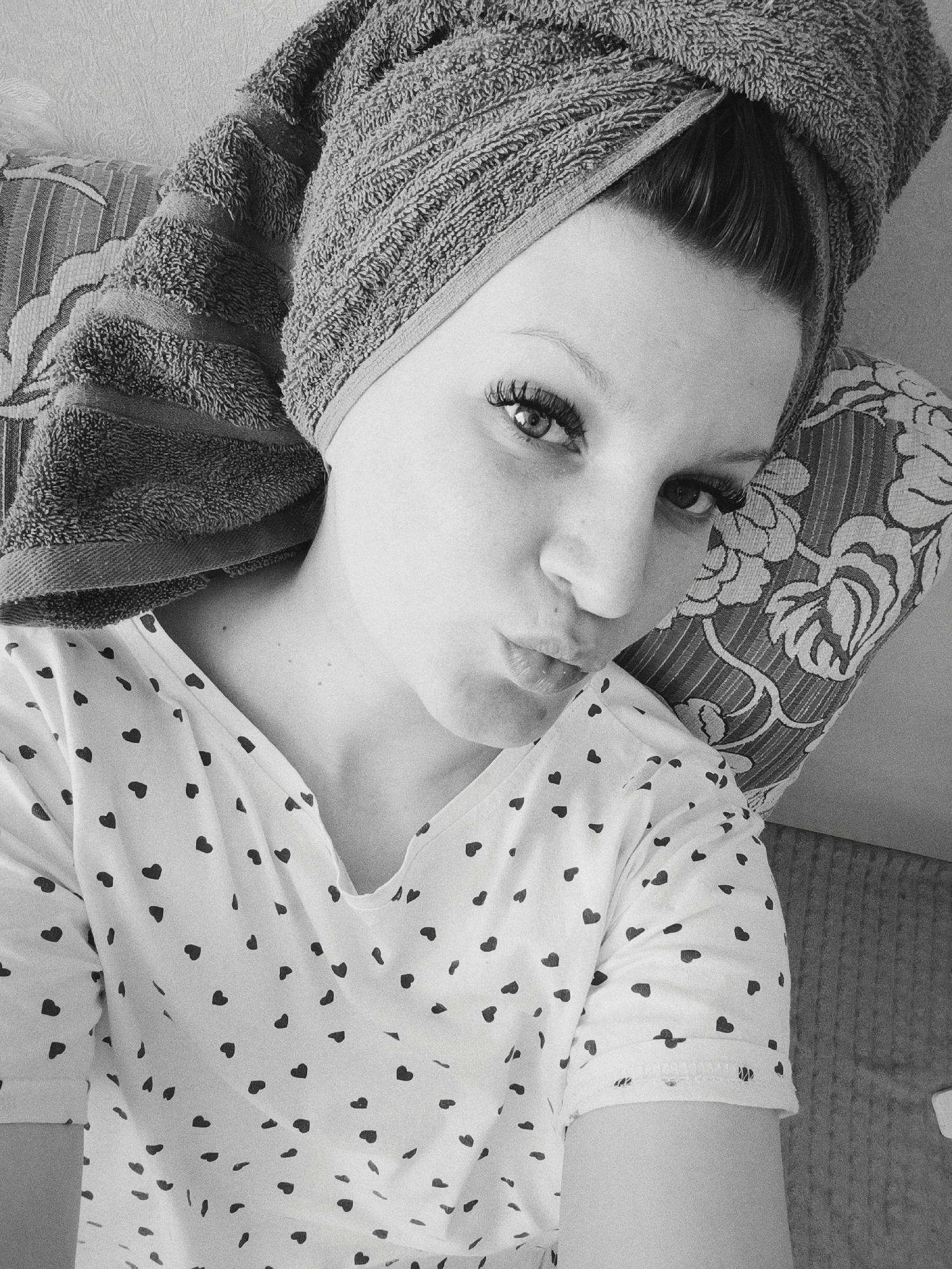 A black and white, close-up, high-angle shot shows a woman with a towel wrapped around her head, puckering her lips in a kiss. She is lying on a floral patterned pillow on a couch. She is wearing a white, v-neck shirt covered in black hearts. Her eyes are open, and she is looking at the camera. The location is presumed to be indoors. The city of Tobolsk, Russia is not visible in the image.