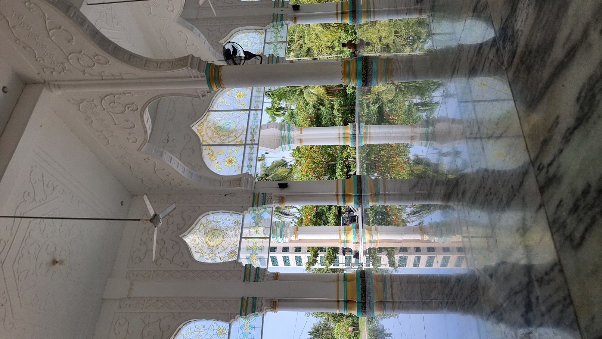 The image depicts the interior of a mosque, specifically the portico area, in Lapang Timu, Indonesia. The architecture features white columns with decorative Islamic motifs and arched openings. Stained-glass windows with floral patterns are visible above the arches. The floor is made of polished marble, creating a highly reflective surface that mirrors the columns and the outdoor scenery. Lush green trees and vegetation can be seen through the openings, suggesting a tropical environment. A distant building with green accents is also visible. In the foreground, the reflection of the architecture dominates the marble floor. To the right, a person is seated on the floor, facing away from the camera, observing the surroundings. A ceiling fan is mounted on the white, ornate ceiling. The overall impression is one of serene grandeur and the integration of natural beauty with religious architecture.