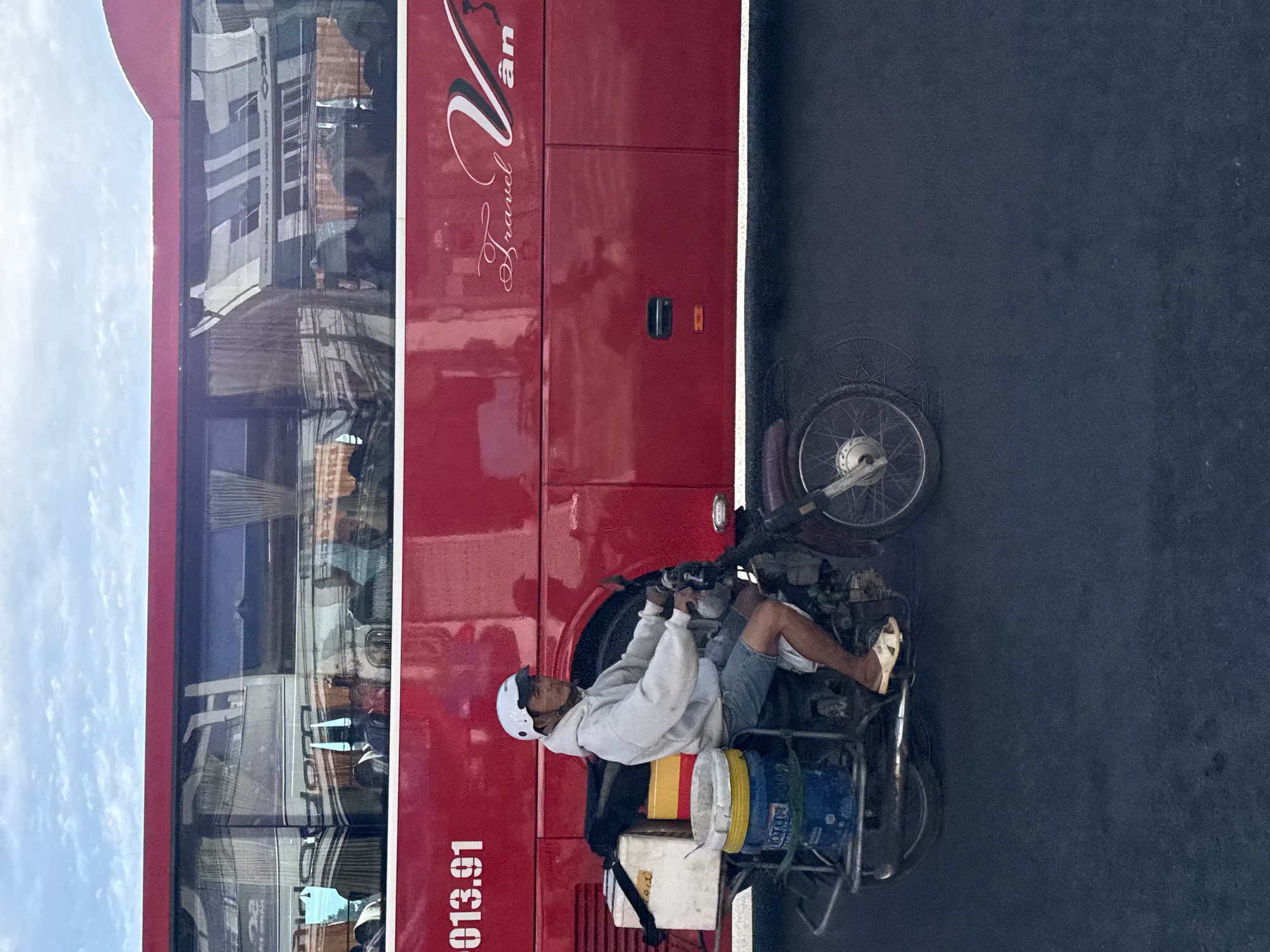 A man wearing a white helmet and a light gray hoodie rides a motorcycle with a sidecar in Da Lat, Vietnam. The motorcycle is loaded with several large plastic containers. The man is wearing denim shorts and beige sandals. In the background, a large red bus is parked or driving alongside the motorcycle. The bus has "Travel Van" written on its side in script. The sky is cloudy.