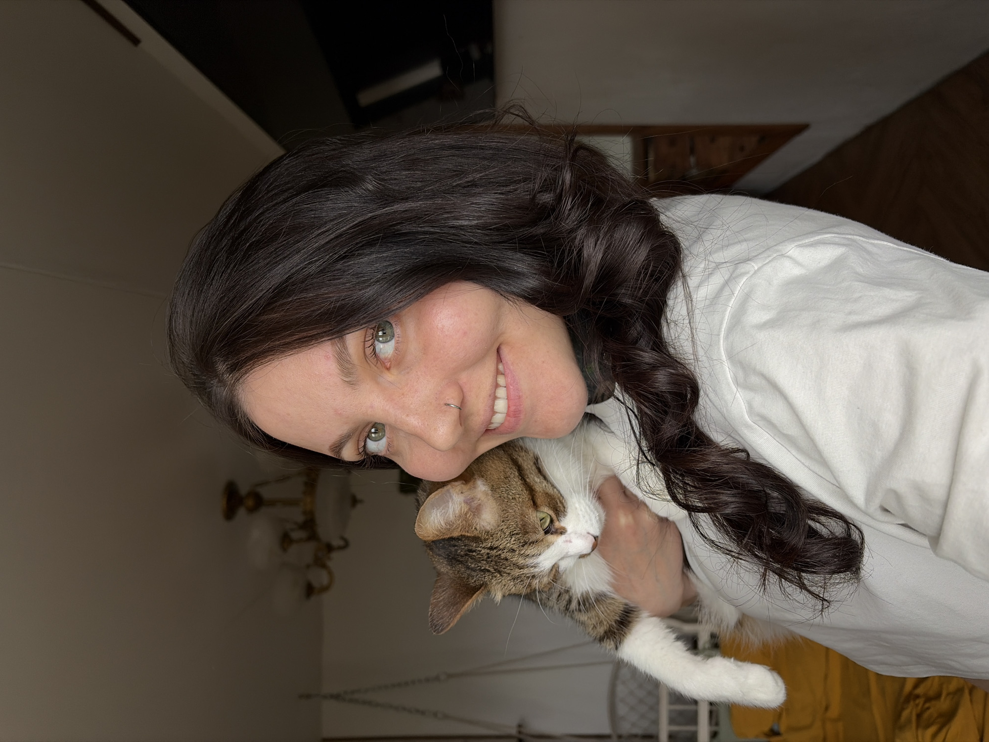 An indoor medium close-up shows a woman holding a cat, tilted counter-clockwise. The woman, positioned on the left side of the frame, has long, dark, wavy hair cascading over her shoulder. She has light green or hazel eyes, a subtle nose piercing on her right nostril, and is smiling broadly, revealing her teeth. She wears a white top.
She holds a domestic short-haired cat, which has a distinctive brown and white tabby pattern with patches of light orange or cream, particularly on its face and body. The cat has green-yellow eyes and prominent white paws. It is held securely in the woman's arms, looking calmly to its right.
The background, appearing somewhat dim, suggests an interior setting, possibly a home in Chaikovsky, Russia. Visible elements include a dark wooden piece of furniture or shelving in the upper background and a brass-colored chandelier with multiple arms and light fixtures in the lower left. The walls are light-colored. The lighting appears soft, indicating an indoor shot, likely during the day. The overall mood conveyed is one of warmth and affection between the woman and her pet. There is no visible text in the image.