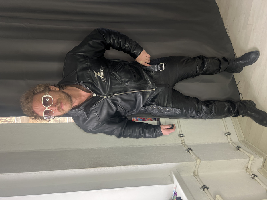 An adult male stands indoors, facing the camera with a slight turn to his left. He has reddish-blonde curly hair, a mustache, and a beard. His eyes are obscured by large, white-framed sunglasses. He is dressed in a black leather jacket featuring a silver zipper, collar, and breast pockets, with a "V-Max" logo and eagle emblem on the left chest. A brown t-shirt is visible beneath the jacket. He wears black leather trousers with decorative straps and a buckle detail on the right hip, paired with black leather boots.
The immediate environment consists of a white-painted wall on the left, featuring a window sill upon which small, colorful items are visible. Below the window, white plastic pipes are mounted horizontally and diagonally along the wall with dark clamps. To the man's right, a dark grey curtain hangs as a backdrop. Through an upper window section, a light-colored, multi-story building with ornate window frames and classical architectural elements is visible, consistent with the architectural styles found in Saint Petersburg, Russia. The floor is a light, neutral color.