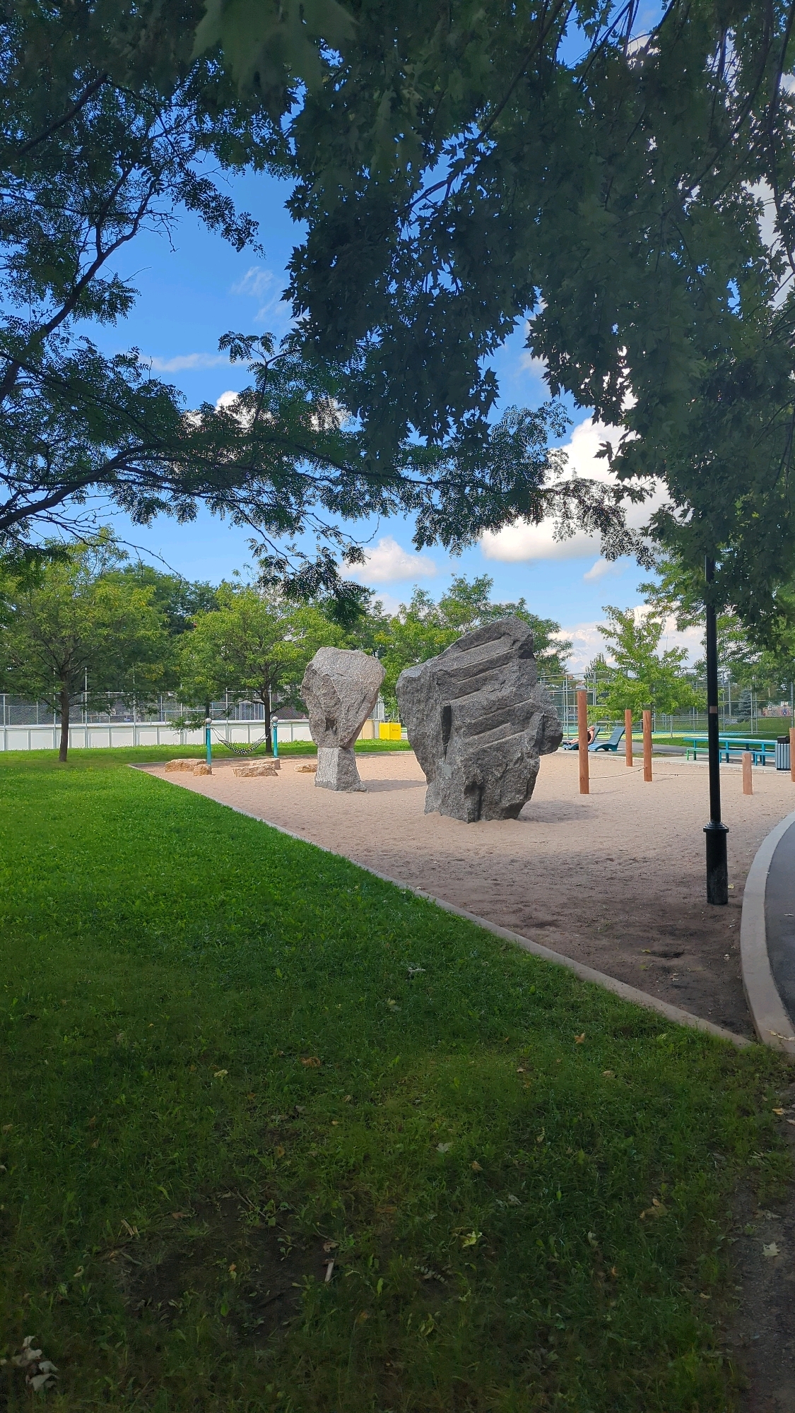 This image captures a section of a public park on a bright, partly cloudy day. The foreground is dominated by a well-maintained grassy lawn with a curved asphalt path bordering its right side. Beyond the lawn, a sand-filled area serves as a playground or recreational space. Two large, grey, sculpted boulders stand prominently within the sand. One boulder is smooth and egg-shaped, resting on a pedestal, while the other is larger, rougher, and features carved lines. In the background, trees with green foliage line the perimeter of the park, and a fence encloses a sports court, possibly a tennis or pickleball court, with a green surface. A few wooden posts and blue seating structures are visible near the sand area. The sky above is a vibrant blue with scattered white clouds. The scene is devoid of any people. Based on the presence of these specific artistic rock sculptures, this location is likely the "Parc des Sept-Roches" in Montreal, Canada, known for its distinctive rock installations.