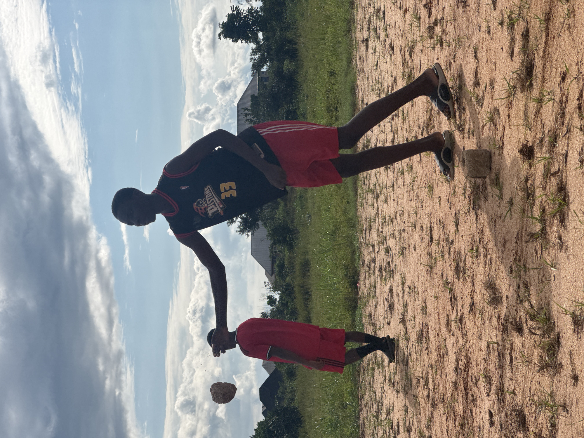 The media file shows two young Black males outdoors on a sandy, uneven surface with sparse grass. One young male, appearing to be in his late teens or early twenties, stands upright. He wears a black basketball jersey with red trim and the number "33" prominently displayed, along with red athletic shorts. His arms are outstretched, with his right hand seemingly in the process of throwing a small, brown, rock-like object. His gaze is directed downward, possibly at the object he is throwing or at a point in the distance.
The second young male is positioned lower in the frame, as if reflected, and is wearing a red jersey with black trim and black shorts. He also appears to be in the process of throwing a similar object. This creates a mirror-like effect where both individuals are engaged in the same action.
The setting includes a background with green vegetation, trees, and what appears to be a building with a pitched roof, suggesting a semi-rural or residential area. The sky is partly cloudy with fluffy white clouds against a bright blue backdrop, indicating daytime with fair weather. The ground is composed of reddish-brown sand and soil, with occasional blades of grass pushing through. The lighting suggests it is a sunny day, possibly late morning or early afternoon, given the shadows cast by the subjects and a small square object near the feet of the upright male. The overall mood seems casual and perhaps playful, given the activity. There is no visible text in the image.