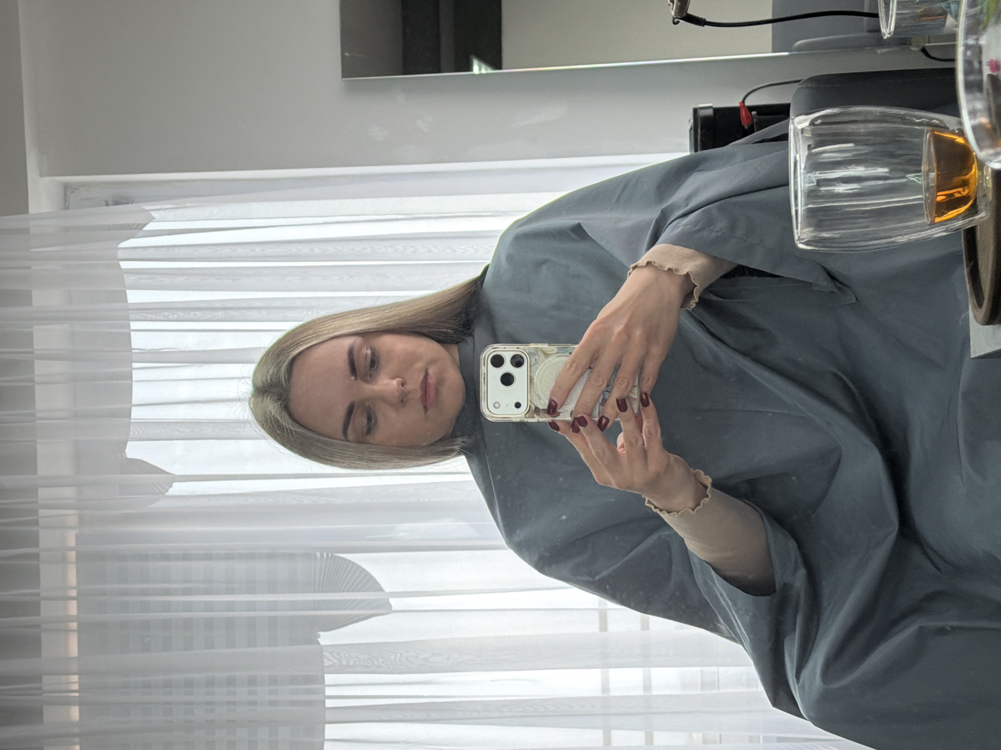 A woman is taking a selfie indoors, wearing a gray smock over a beige shirt with ruffled sleeves. She is holding a phone in her hands with dark red nail polish. A glass with a yellow liquid is to her right. The location is not identifiable as Votkinsk, Russia, based solely on the given image. Background includes white curtains and a mirror reflecting part of the room.