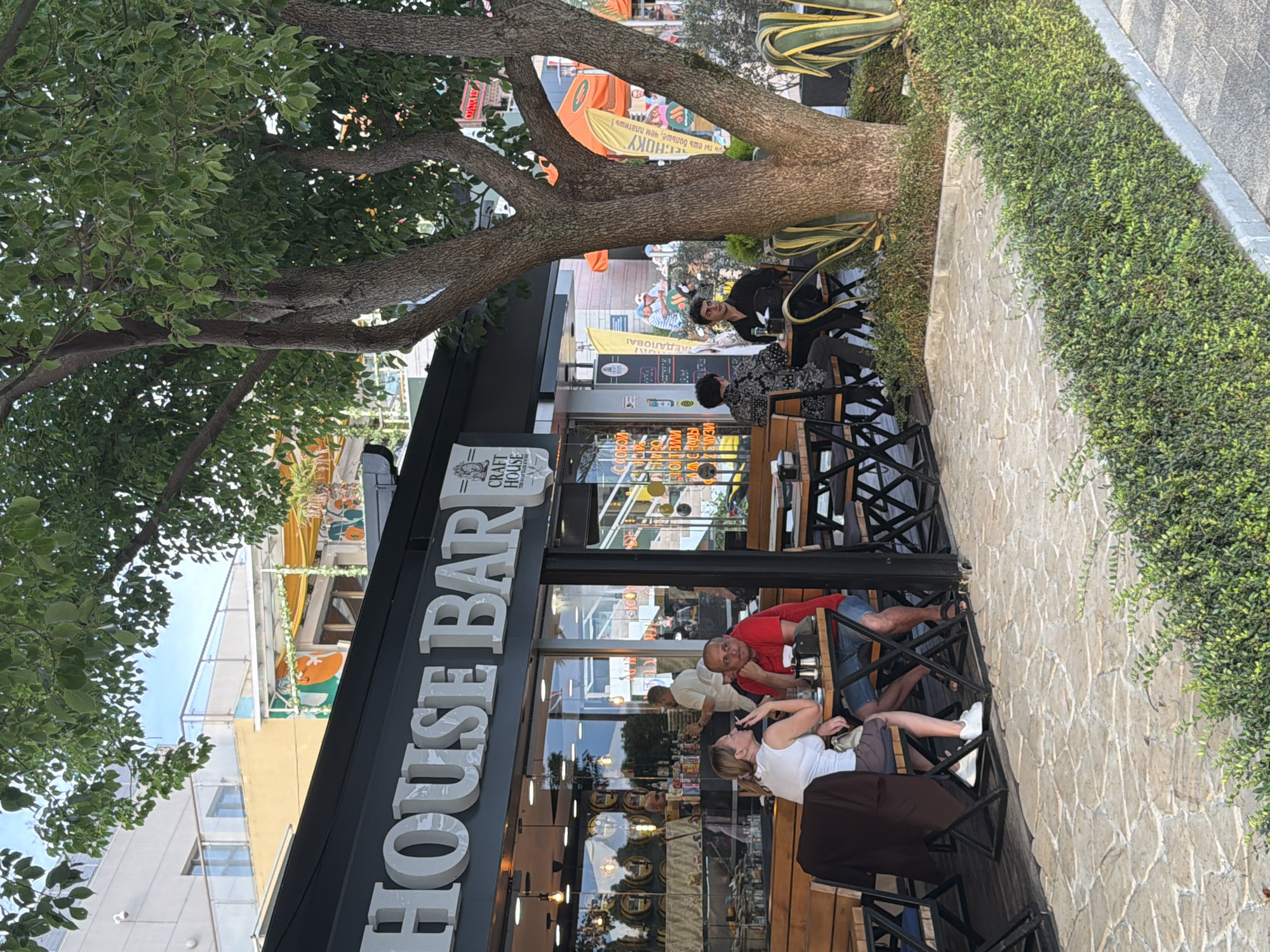This image depicts an outdoor seating area of a establishment identified by the sign "CRAFT HOUSE BAR". The establishment appears to be located in Sochi, Russia, as indicated by the Cyrillic text visible on signs and promotional materials within the scene, specifically phrases like "С СОБОЙ" (with you/for takeaway) and what appears to be a Russian slogan on a yellow banner.
In the foreground, a couple is seated at a table. The woman on the left, wearing a white top and a brown skirt, is leaning forward and appears to be talking. The man on the right, wearing a red polo shirt and denim shorts, is looking towards the woman. Next to them, on a table, are salt and pepper shakers, a cellphone, and a bottle.
Behind them, other patrons are visible. A man in a white shirt is standing inside the bar area, possibly attending to customers. Further back, a person in a black shirt is seated at a table. To the right, a large tree with dense foliage shades the outdoor seating area.
The establishment features a modern design with large glass windows, allowing a view into the interior where barrels and bottles are displayed. The outdoor seating area is paved with stone tiles and bordered by a manicured hedge. Various banners and signs with Cyrillic text are visible, contributing to the specific cultural and geographical context of the location. The overall atmosphere suggests a relaxed daytime setting at a bar or cafe.