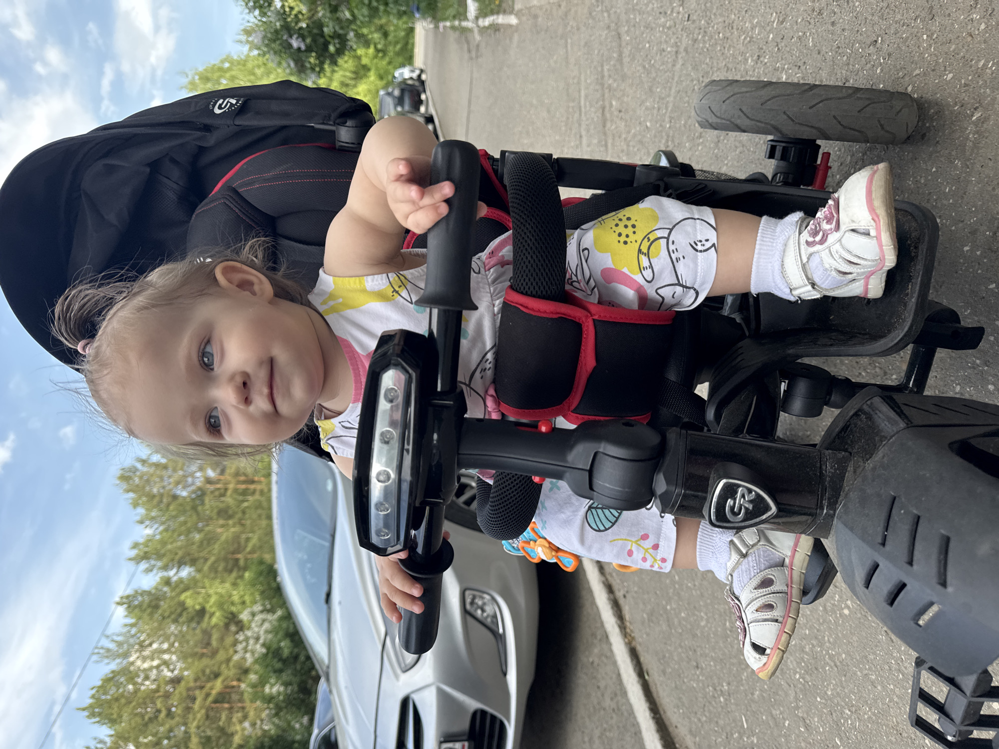 A young child sits in a tricycle on a paved surface. The tricycle is black with red accents and has a large black canopy. The child is wearing a white and yellow patterned dress, white socks, and white sandals. The tricycle is positioned near a silver car parked on the side of a road. Trees and a building can be seen in the background. The city of Chaikovsky, Russia, cannot be determined from the image.