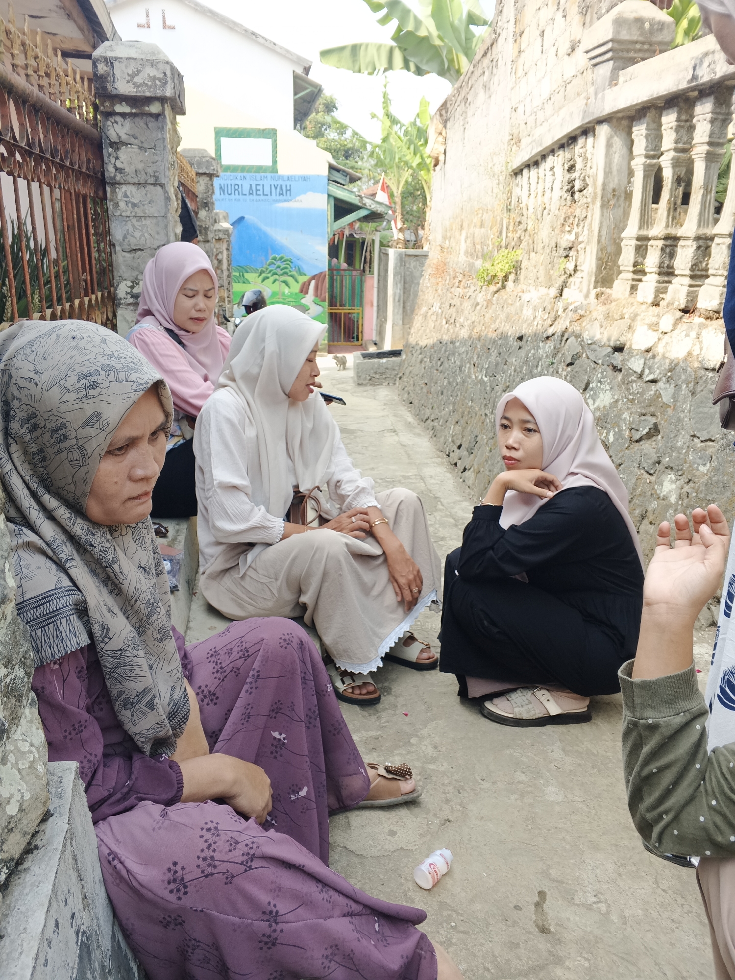 The image shows a group of four women sitting on stone steps outdoors, likely in front of a building in Warungkiara, Indonesia. The building behind them has a mural painted on its wall that reads "PENDIDIKAN ISLAM NUR LAELIYAH" and depicts a mountainous landscape with a waterfall. The women are all wearing hijabs, with varying colors and patterns. The woman in the foreground on the left is wearing a purple dress with a floral pattern and a grey hijab with an intricate design. To her right, another woman in a light-colored hijab and a beige outfit is seated. Further to the right, a woman in a black outfit and a light pink hijab is crouched down, resting her chin on her hand. A fourth woman, partially visible on the far right, is wearing a green shirt with white polka dots and appears to be speaking or interacting with the woman in the black outfit. A small bottle of yogurt drink is on the ground near the women. The environment appears to be a street or pathway, with stone walls and some greenery visible. The lighting suggests it is daytime.