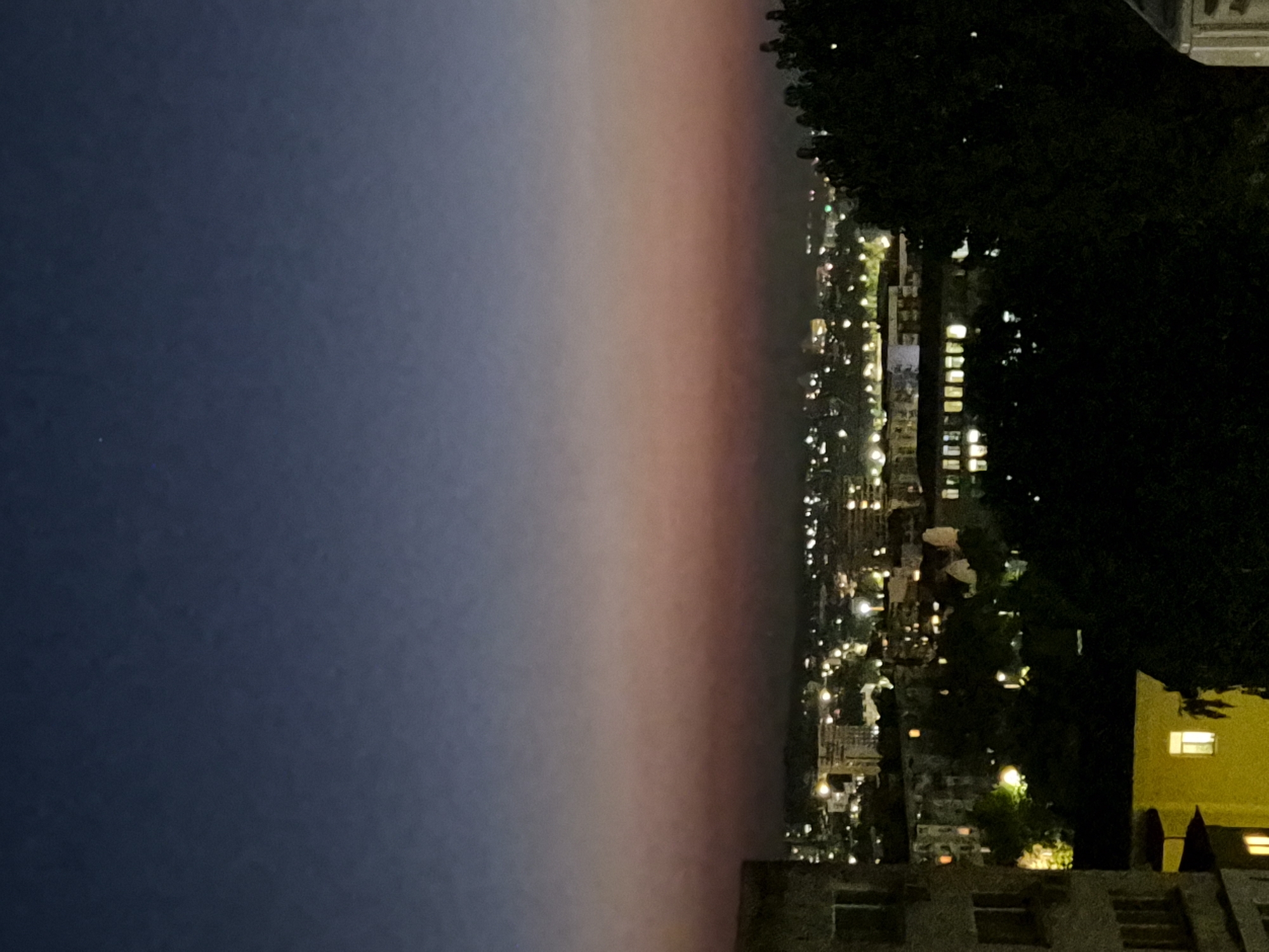 The image depicts a cityscape at dusk, as indicated by the gradient of the sky transitioning from deep blue at the top to a soft pink and orange hue closer to the horizon. The city lights are illuminated, creating a warm glow against the darkening sky. In the foreground, a dark silhouette of trees partially obscures the view of the buildings and lights. To the left, a portion of a light-colored building is visible, showing windows and architectural details. Further into the city, numerous buildings of varying sizes and shapes are scattered, with many windows lit. The overall scene suggests an urban environment at the end of the day, with the transition from daylight to night.
