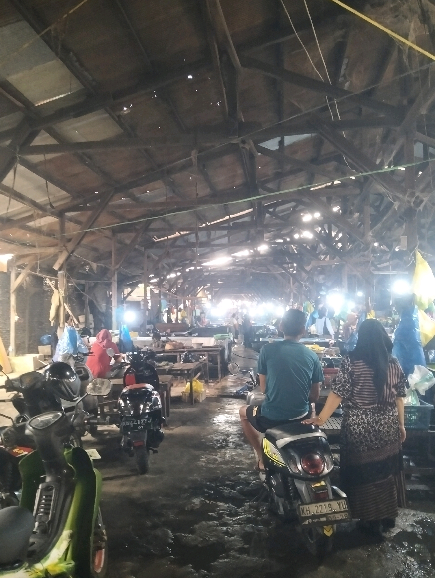 The image depicts an interior view of an open-air market, likely a traditional wet market, in Palangka Raya, Indonesia. The structure appears aged, with a prominent wooden beam ceiling showing signs of wear and tear, and some translucent panels allowing light to filter through. The ground is uneven and wet, suggesting recent rain or a constant presence of water.
In the foreground, several motorbikes are parked. One is a green scooter on the left, partially visible. To its right, a black scooter with a visible license plate (KH 2219 YU) is occupied by a person wearing a teal t-shirt and black shorts. A woman with long dark hair, wearing a patterned dress, stands next to the scooter, her hand resting on the seat. In the middle ground, a person in a red headscarf is seated on a motorbike.
The market is filled with various stalls and activity. Several tables are set up, some with goods displayed. In the background, more people can be seen browsing or working. Bright lights illuminate different sections of the market, creating a hazy, atmospheric effect. Colorful tarps, possibly blue and yellow, are visible hanging above some stalls. The overall impression is of a bustling, functional marketplace in an outdoor setting.