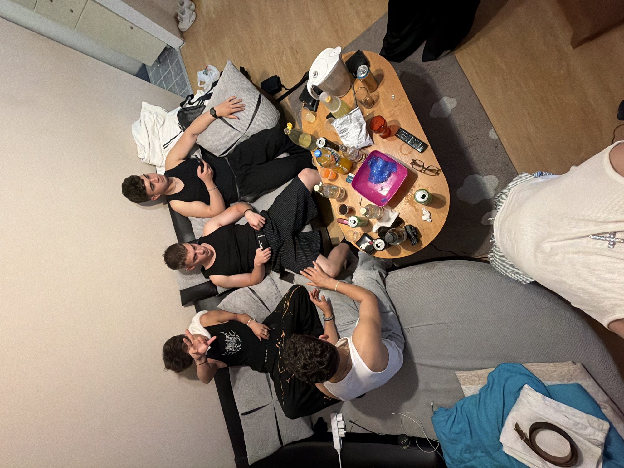 Four young men are lounging on a grey sofa. A coffee table is positioned nearby, cluttered with bottles, cans, glasses, a remote control, and a pair of eyeglasses. The location is not discernible from the image.