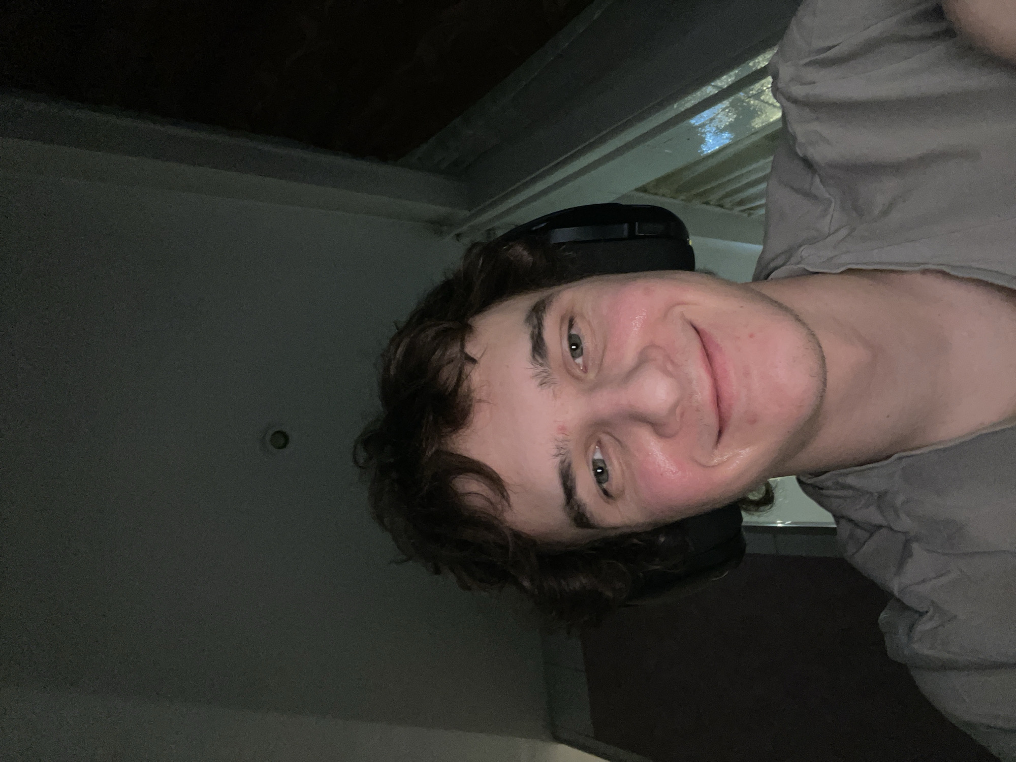 A selfie of a young Caucasian man with dark curly hair and a headset is illuminated by a light source above. The man is smiling with moist skin. He is wearing a gray t-shirt. The background shows a dark ceiling and a light fixture. The location is indeterminate, and there is no information available to suggest this image was taken in Krasnoyarsk, Russia.