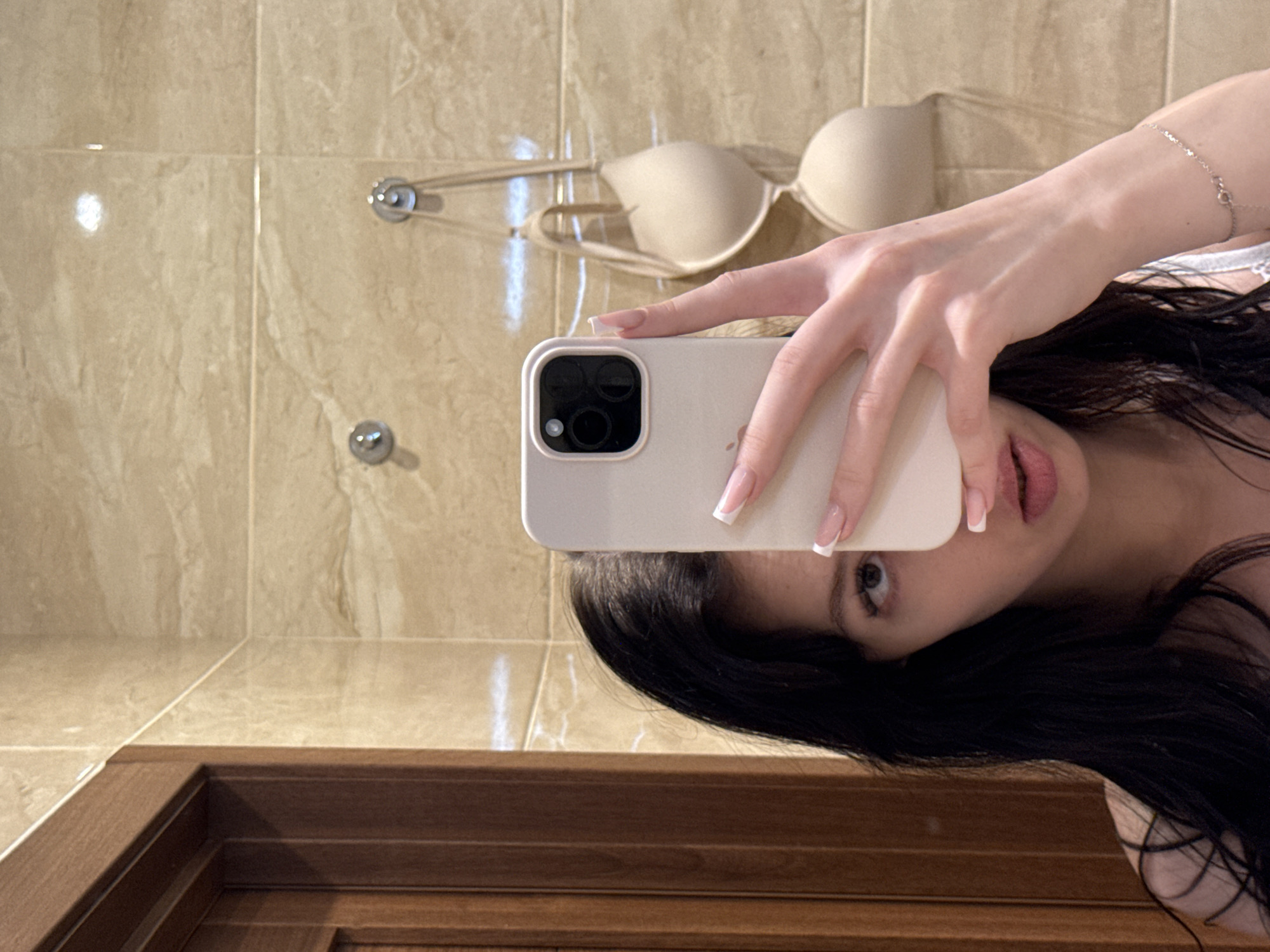 A young woman with long, dark hair takes a selfie in a bathroom with beige tiled walls and a wooden door frame. She is holding a light-colored smartphone in her right hand, partially obscuring her face. A light beige bra is hanging on hooks on the wall behind her. A delicate bracelet is visible on her left wrist. The overall scene suggests a private setting, possibly a home bathroom, within the city of Khimki, Russia.