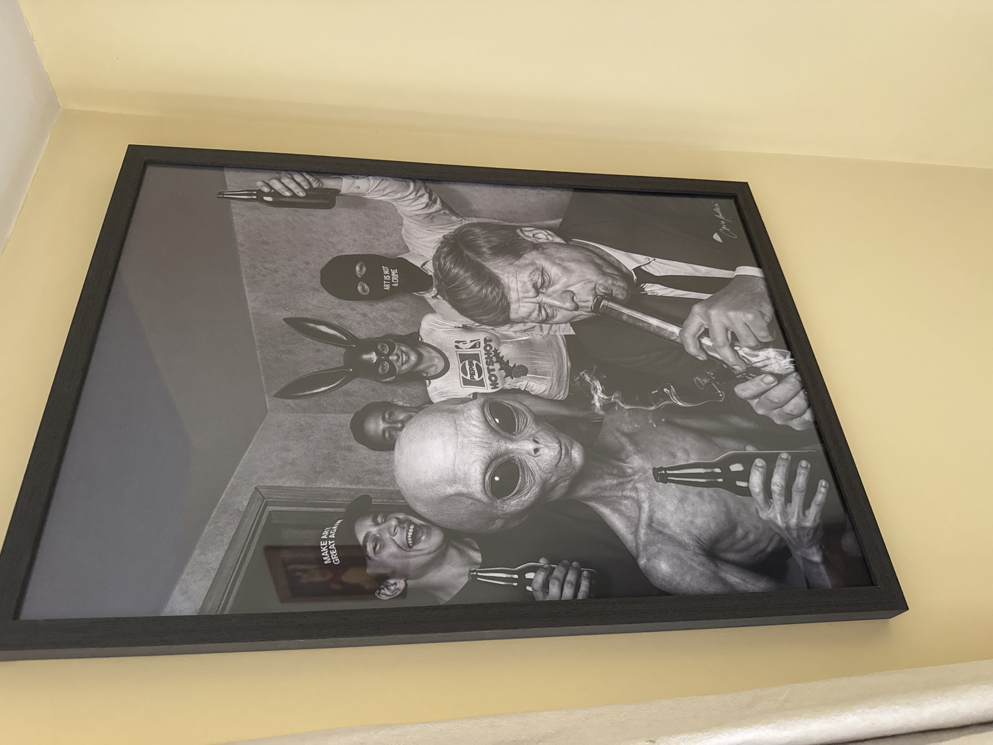 The image displays a framed black and white artwork hanging on a pale yellow wall. The artwork itself depicts a surreal scene with several figures. In the foreground, John F. Kennedy is shown smoking from a pipe. To his left, an alien figure holds a bottle. Behind them, a group of individuals are partially visible; one wears a ski mask with "ART IS NOT A CRIME" printed on it, another wears bunny ears, and a third person is laughing while wearing a cap that reads "MAKE ART GREAT AGAIN." The artwork is positioned to the right of a white curtain. The image is a photograph of this artwork. There is no information to suggest this is located in Colchester, United Kingdom.