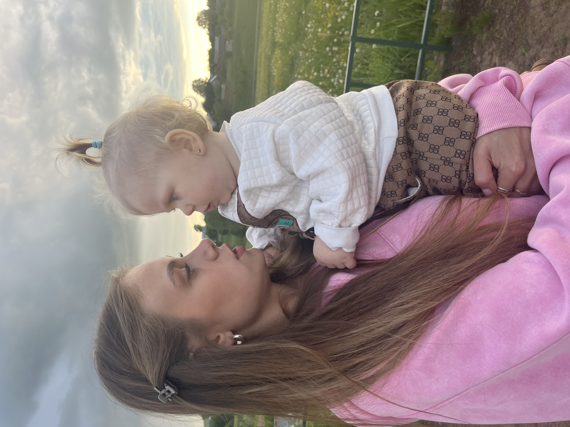 A woman in a pink sweatshirt lies on her back in a grassy field, holding a baby on her stomach. The baby wears a white quilted sweatshirt and brown monogrammed overalls. The woman and baby are looking at each other. The sky is overcast with the sun setting on the horizon, and a small building is visible in the background. The location is Фоки, Russia.