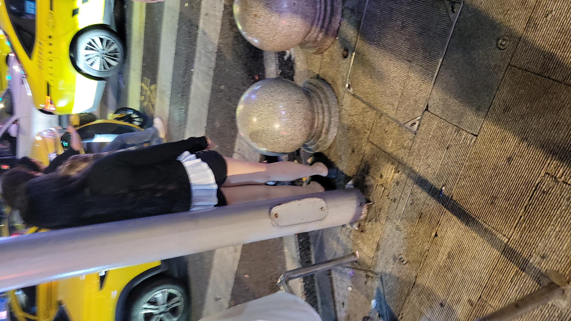 The image depicts a scene in an urban environment, seemingly at night, with multiple elements:
**People:** There is a person lying horizontally near what appears to be a light pole. They are wearing dark attire with what appears to be a light-colored trim or skirt. Additionally, there's at least one other person standing in the background.
**Objects:** The scene includes street elements such as light poles topped with spherical lights. There are also vehicles, specifically yellow cars or taxis. The environment appears to be a street or pedestrian area, and there is evidence of infrastructure such as pavement.
**Location:** The image does not provide sufficient detail to ascertain the specific city or location.
**Environment:** The setting is urban and appears to be in the evening or at night, given the lighting conditions.
**Interactions:** The person is lying prone on the ground, seemingly interacting with the light pole. The image does not reveal their condition or any other interactions.