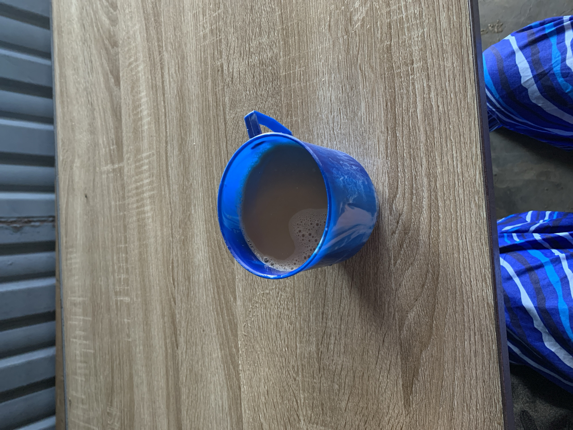 A blue plastic mug filled with a tan-colored liquid sits on a wooden surface. The surface appears to be a tabletop. To the left of the table is a corrugated metal wall. To the right is a patterned blue and white fabric. The scene is likely indoors. The location is Rafin Par, Nigeria. There are no people or interactions visible.