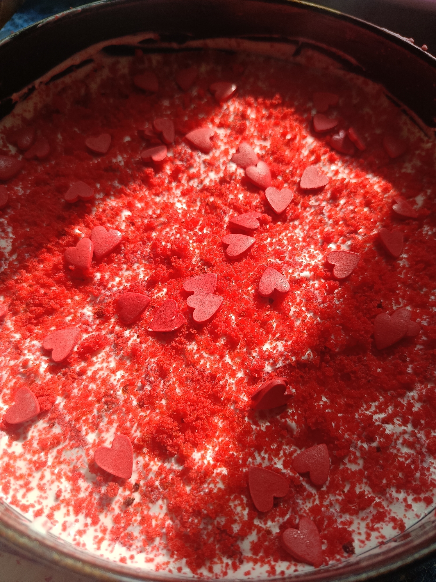 A close-up, top-down view captures a dessert, likely a cake or cheesecake, prepared in a dark, round baking pan. The surface of the dessert is a creamy white or light-colored base, heavily adorned with vibrant red toppings. These toppings consist of a dense layer of fine, granular red sprinkles, resembling crushed red velvet cake, distributed across the dessert's surface. Interspersed among the red sprinkles are numerous distinct, smooth, red heart-shaped candies or sprinkles.
Natural light, possibly sunlight, illuminates the scene from an angle, creating bright highlights on the heart shapes and casting subtle shadows, suggesting the photograph was taken during daytime. The contrasting rich red and creamy white colors evoke a sense of sweetness and celebration, strongly implying a romantic theme, such as for Valentine's Day or an anniversary. The texture appears to be a mix of the soft, creamy base, finely crushed red crumbs, and smooth heart candies. No specific activity is depicted; the image showcases the finished, decorated dessert. There is no visible text within the image. The location context provided is Rusalevka, Russia.