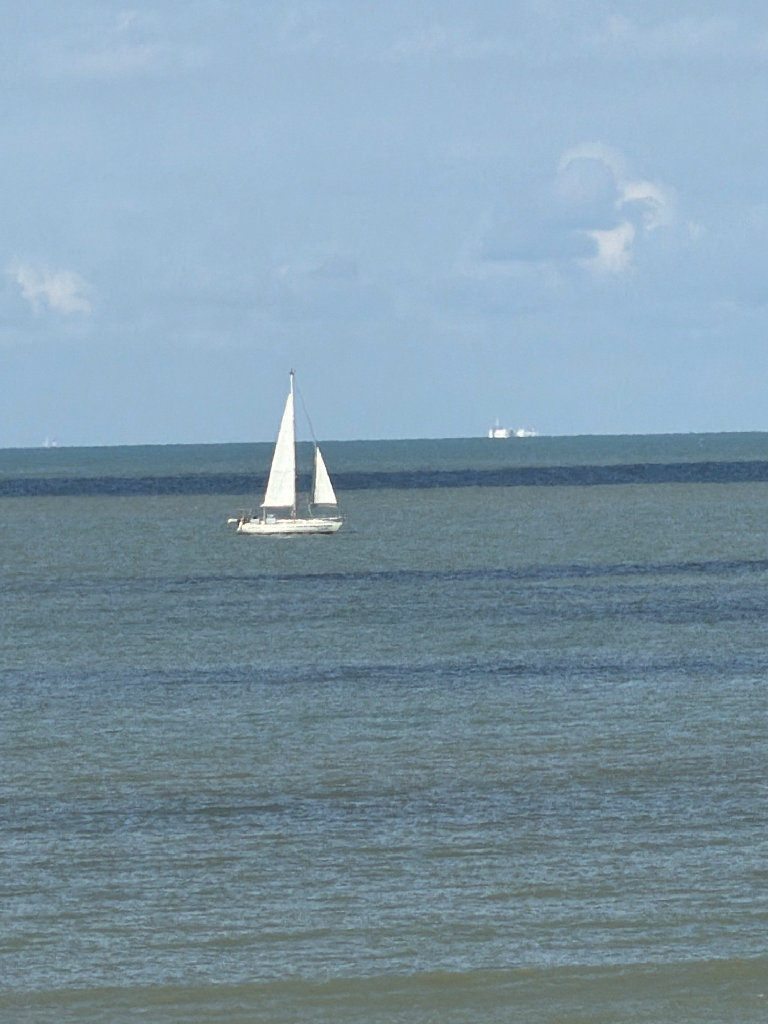 The image shows a sailboat with white sails on a calm sea under a cloudy sky. In the distance, a large ship or oil rig is visible on the horizon. The scene appears to be offshore from Ostend, Belgium. The water is a deep blue-green, and the sky is a pale blue with scattered white clouds. The sailboat is the main subject of the image, and it is positioned in the center-left of the frame. The large distant object is on the horizon line, to the right of the sailboat.