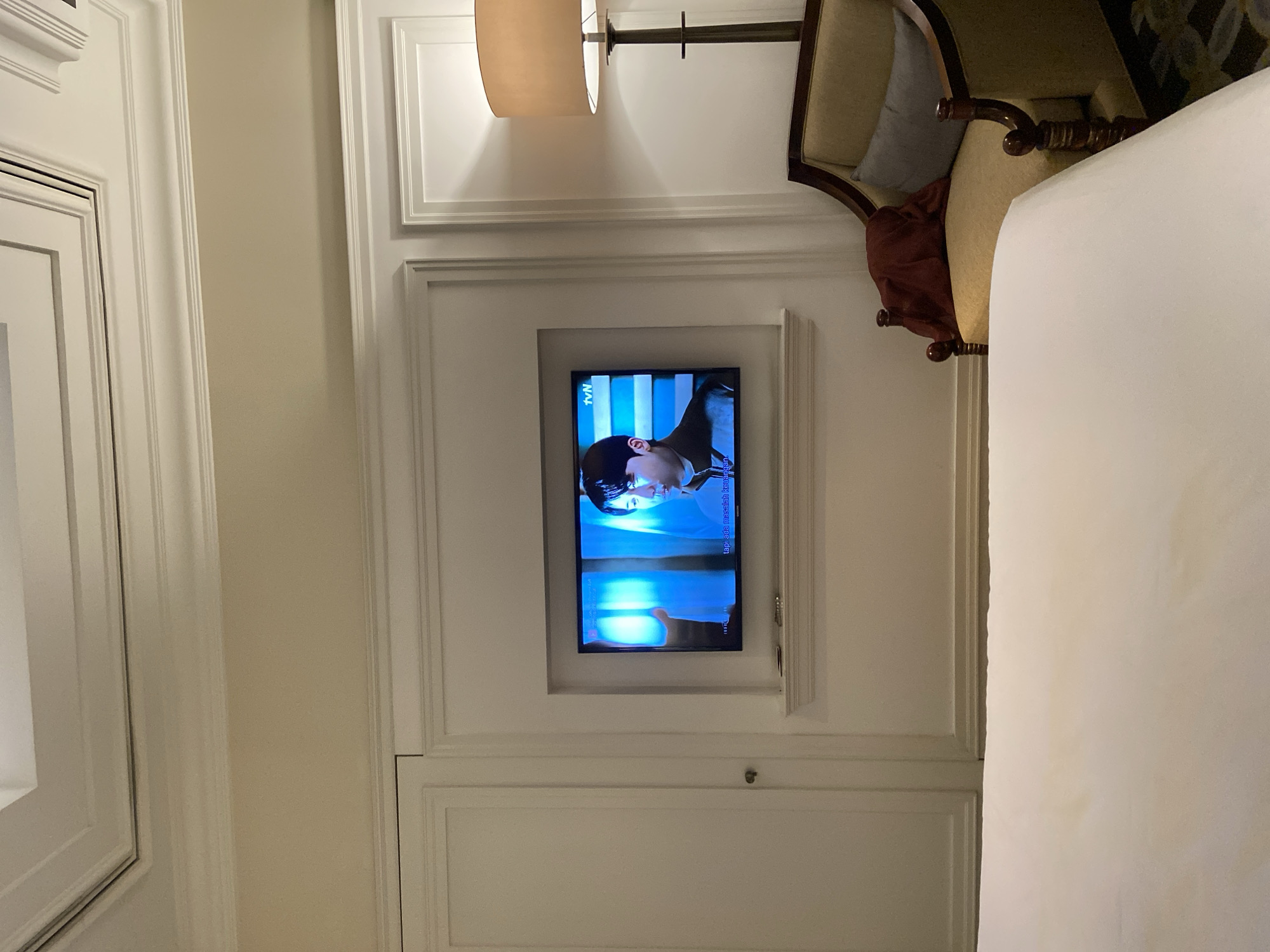 This image shows a bedroom with a TV mounted on a wall. The TV displays a scene from a show, with the "tvN" logo visible in the top left corner. The room is decorated with white paneled walls and a beige wall. There is a lamp with a beige shade hanging from the ceiling. To the right of the TV, there is a chair with a light brown seat and dark brown trim, a blue pillow, and a burgundy throw pillow. The overall setting suggests a hotel room or a well-decorated home. The lighting in the room appears to be artificial, possibly from the lamp and the TV screen. The image is taken from a slightly elevated perspective, looking down towards the TV.