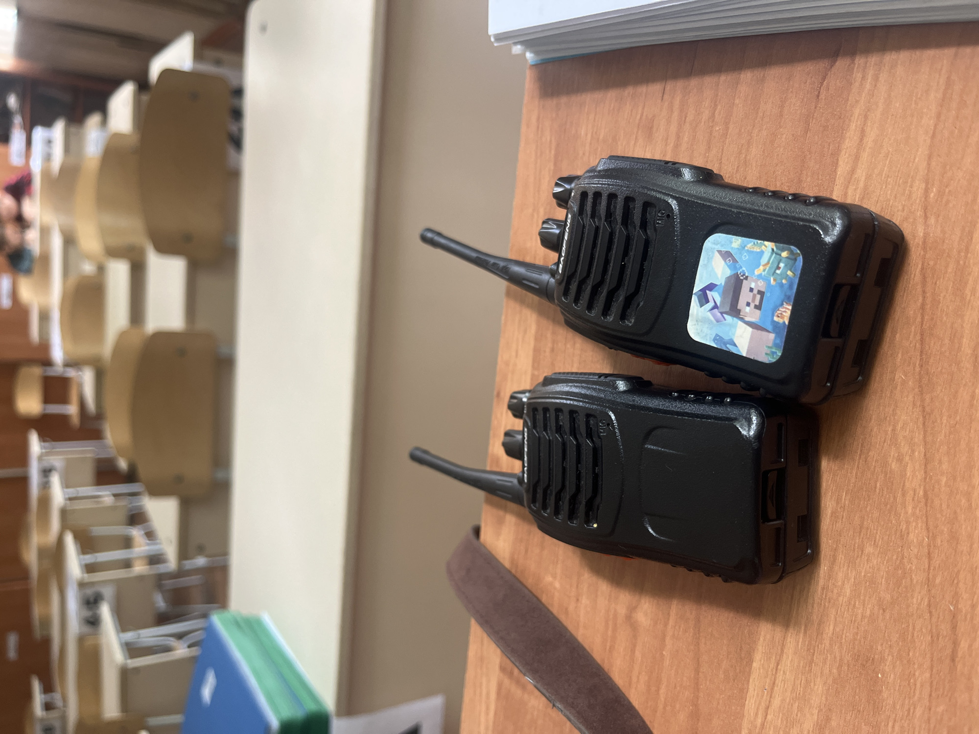 Two black walkie-talkies are placed on a light brown wooden desk in what appears to be a classroom or lecture hall setting in Kaspiysk, Russia. The walkie-talkies are oriented horizontally, with their antennas pointing to the left. The top walkie-talkie features a colorful square sticker depicting characters and structures from the video game Minecraft. Faintly visible on the speaker grille of this walkie-talkie is the brand name "BAOFENG." The bottom walkie-talkie is plain black.
In the blurred background, rows of empty, light-colored chairs and desks are arranged, characteristic of an educational institution. A white vertical partition or wall separates the foreground desk from the background. On the far left, a very blurry human figure or part of one can be seen among the chairs. To the left of the walkie-talkies, a stack of white papers or books is visible. Below the walkie-talkies, a blue and green book or folder rests on the desk. A brown strap, possibly from a bag or belt, is also partially visible on the wooden surface near the bottom of the frame. The overall scene suggests an indoor environment, possibly after school hours or during a break. No specific activity is actively taking place, but the items suggest recent use or preparation for use.
