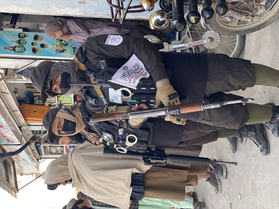 This image depicts an outdoor urban street scene in Kandahar, Afghanistan, likely a commercial district. In the foreground, two armed men stand on a paved surface, both wearing dark long-sleeved uniforms, dark tactical vests, dark baseball caps, and black face masks (N95-style respirators) over balaclava-style head coverings.
The individual on the right holds an AK-pattern rifle with a wooden foregrip and buttstock. He wears tan tactical gloves, and a small white flag with black Arabic script (the flag of the Islamic Emirate of Afghanistan) is affixed to his tactical vest. A radio and pens are also visible on his vest, along with silver-colored handcuffs.
The individual to his left, similarly dressed and equipped, has an M4/AR-15-pattern rifle slung on his left side. Silver-colored handcuffs are also visible on his vest. Both men wear dark knee-length trousers over olive-green socks and dark tactical boots.
To the left of these armed individuals, several adult males in traditional Afghan attire, including turbans, light-colored shawls (patoo), and shalwar kameez, are standing. In the mid-ground right, a young female figure in a light purple dress and head covering stands at the entrance of a shop.
The background consists of commercial shop facades displaying various items, including lighting fixtures and what appear to be auto parts. An awning extends over the shops. Partially visible on the far right is a white motorcycle with chrome elements, and the front wheel and suspension of another motorcycle are in the middle right. The scene is illuminated by natural light, indicating daytime.