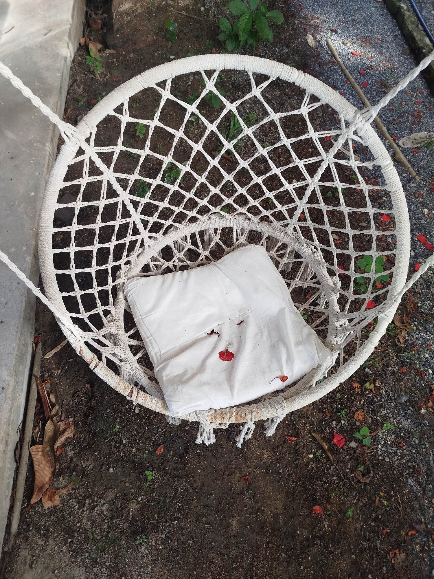 An overhead shot shows a white macrame net chair with a white cushion placed inside. The chair is suspended by ropes from unseen anchor points. The chair and cushion are resting on dark soil with small pebbles and scattered red flower petals, indicative of an outdoor environment. A concrete structure is visible to the left of the chair, and dry leaves are present on the ground. The scene appears to be located in Baan Lamai, Thailand.