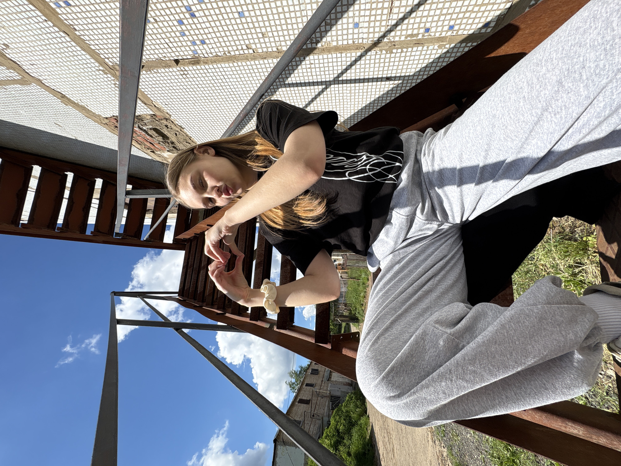 The image presents a young woman sitting on a metal structure, possibly a staircase or a platform, against a backdrop of a blue sky with scattered clouds. The scene is located in Chaikovsky, Russia.
The woman is wearing a black t-shirt with a white graphic design, paired with light grey sweatpants. She is angled towards the viewer and her hands are positioned in front of her, forming a heart shape. A scrunchie adorns her wrist.
The metal structure appears weathered and possibly old. The surrounding environment includes a view of buildings and greenery. The photograph is taken from a low angle, emphasizing the height and structure of the metal platform.